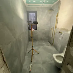 Łazienka w trakcie remontu: szare płytki na ścianach i podłodze, częściowo wykończony sufit z płyt karton gips, złota armatura, oświetlenie robocze na statywie, widoczny fragment toalety...