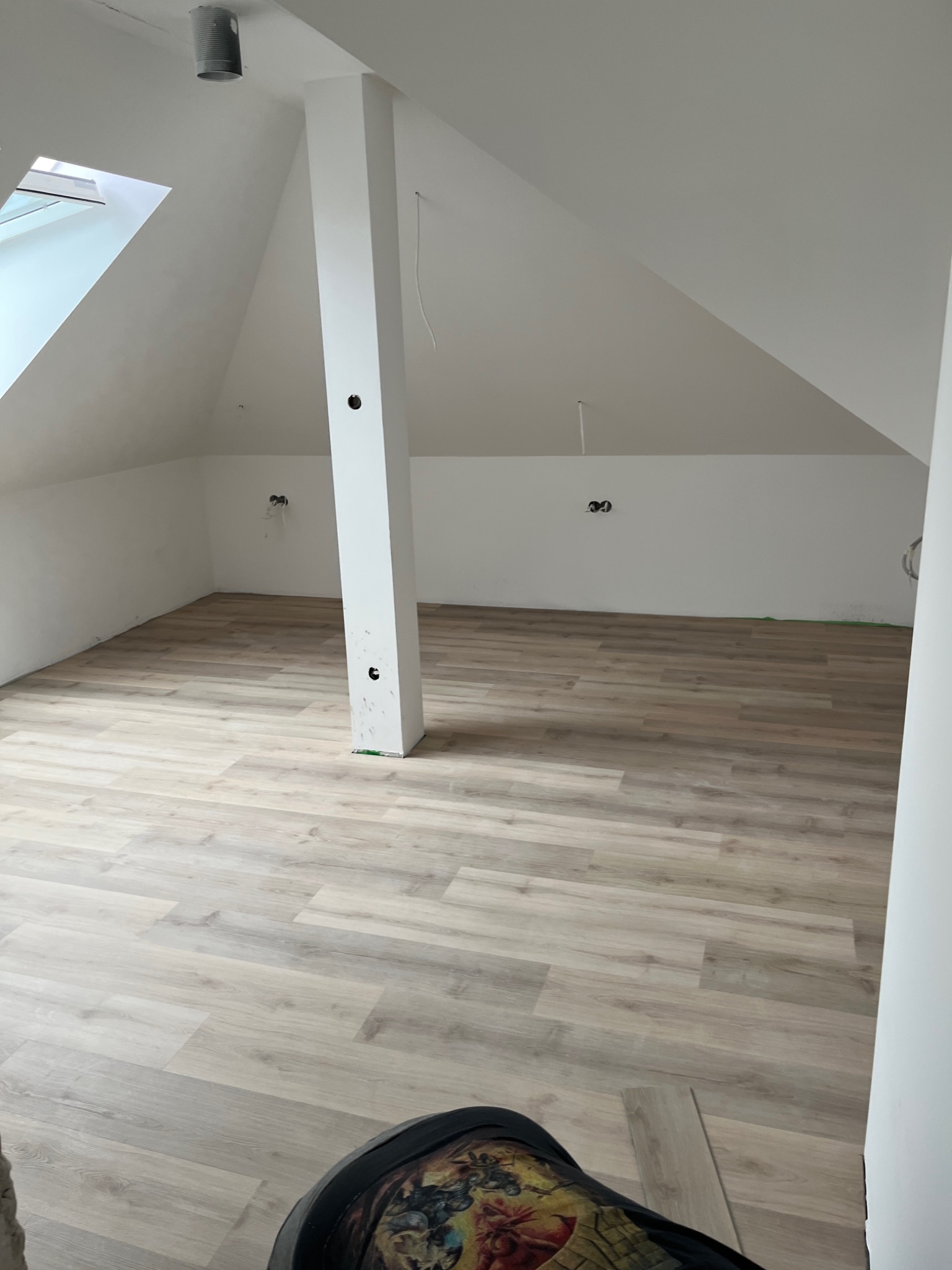 Poddasze z nowo ułożoną podłogą z paneli w odcieniach szarości, widoczny słup konstrukcyjny, okno dachowe i przygotowane punkty elektryczne na ścianie.