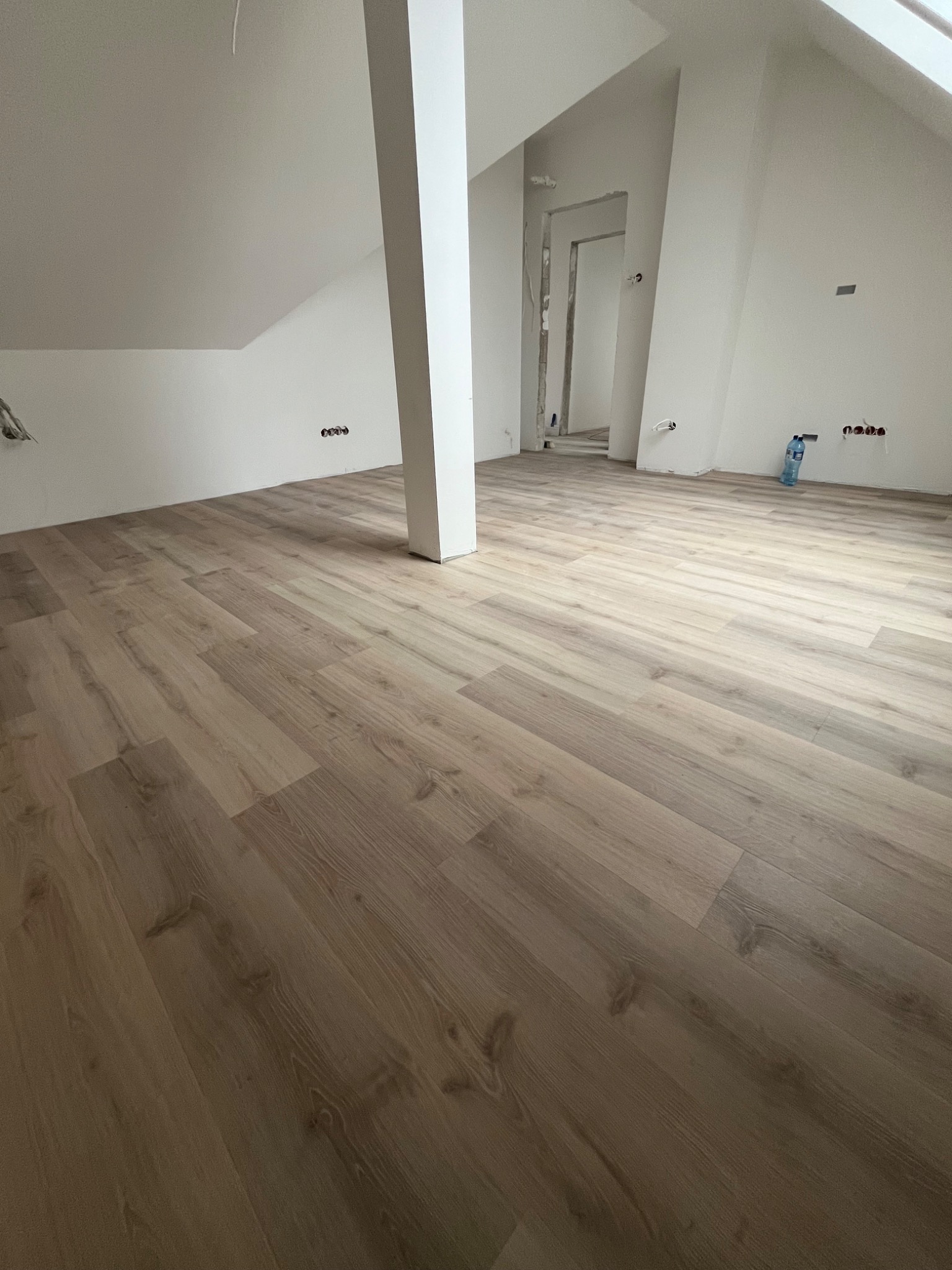 Jasne panele podłogowe ułożone w pokoju na poddaszu z białymi ścianami i słupem nośnym, widoczne gniazdka elektryczne i butelka wody mineralnej.