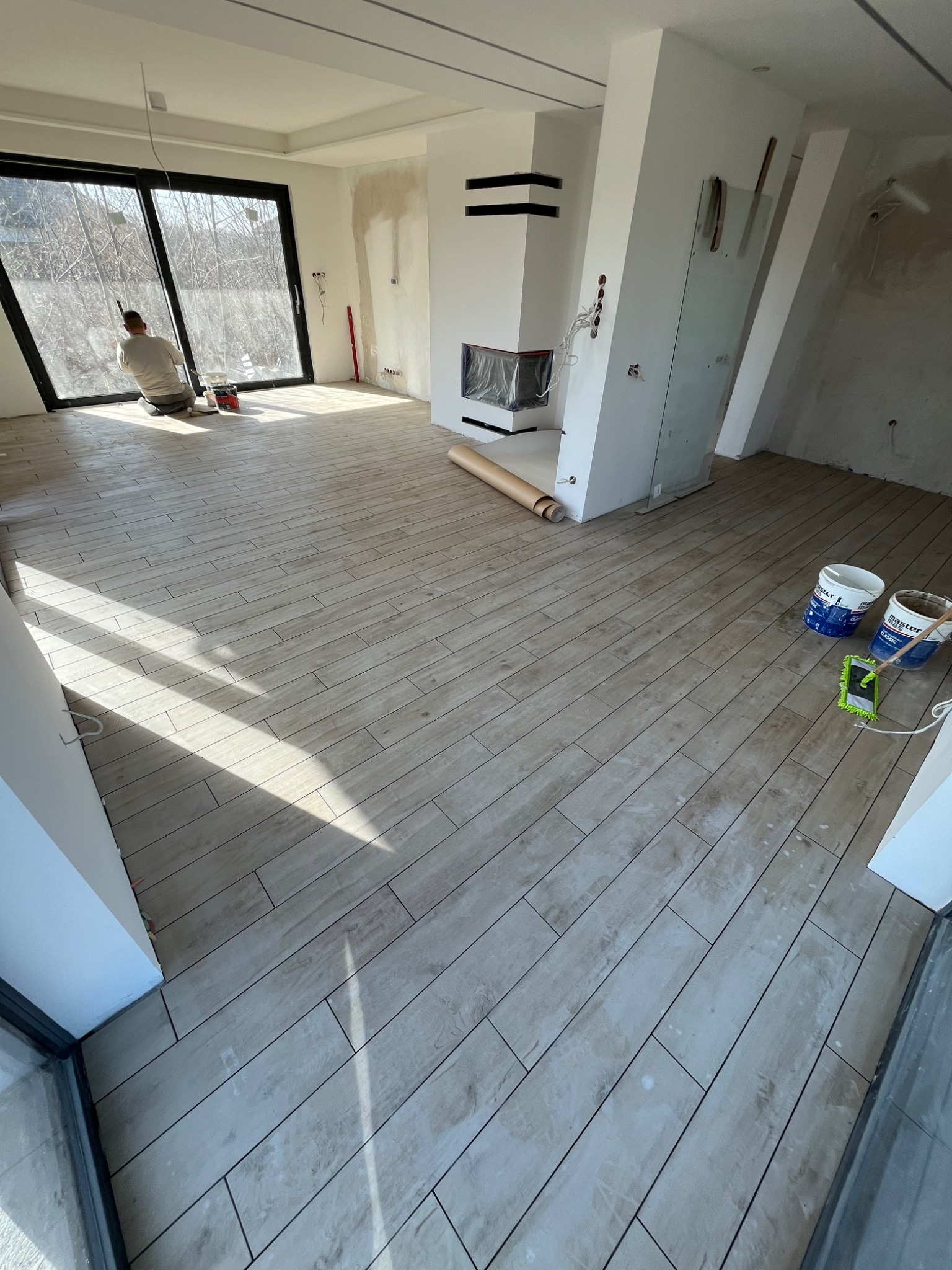 Układanie podłogi z płytek imitujących drewno w przestronnym, jasnym salonie z kominkiem; widoczny pracownik przy oknie, wiaderka z farbą i narzędzia.