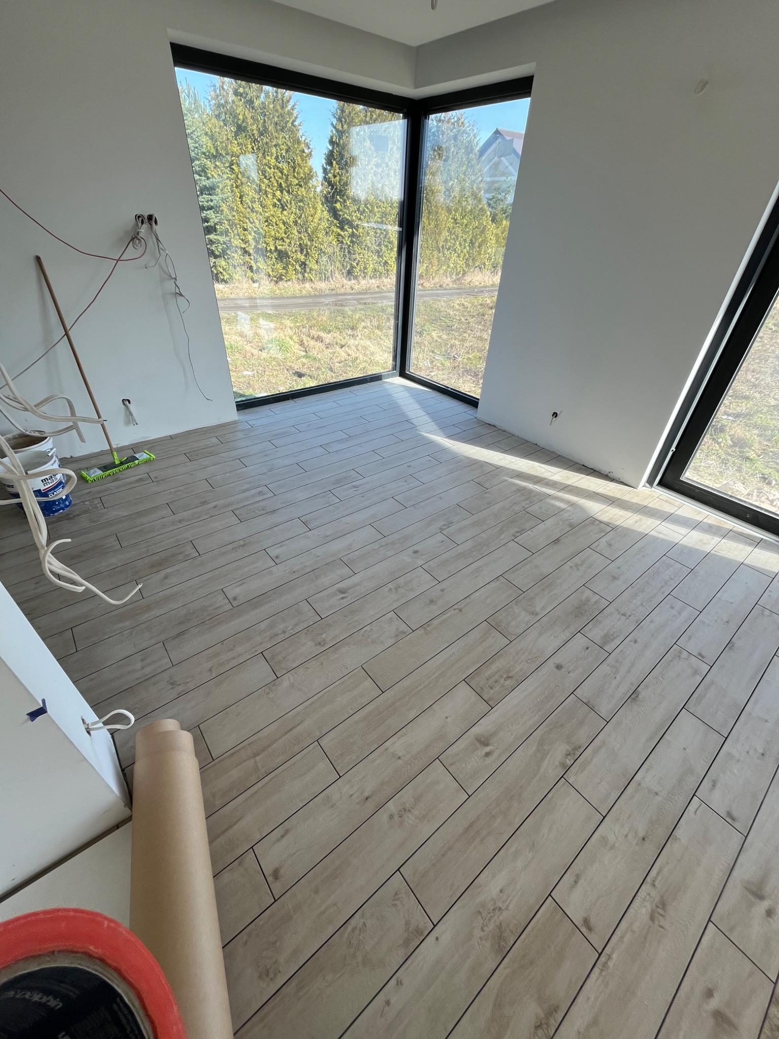 Ułożona podłoga z płytek imitujących drewno w jasnym, nowoczesnym wnętrzu z dużymi oknami. Widoczne kable wystające ze ściany, wiadro farby i wałek malarski sugerują prace wykończeniowe.