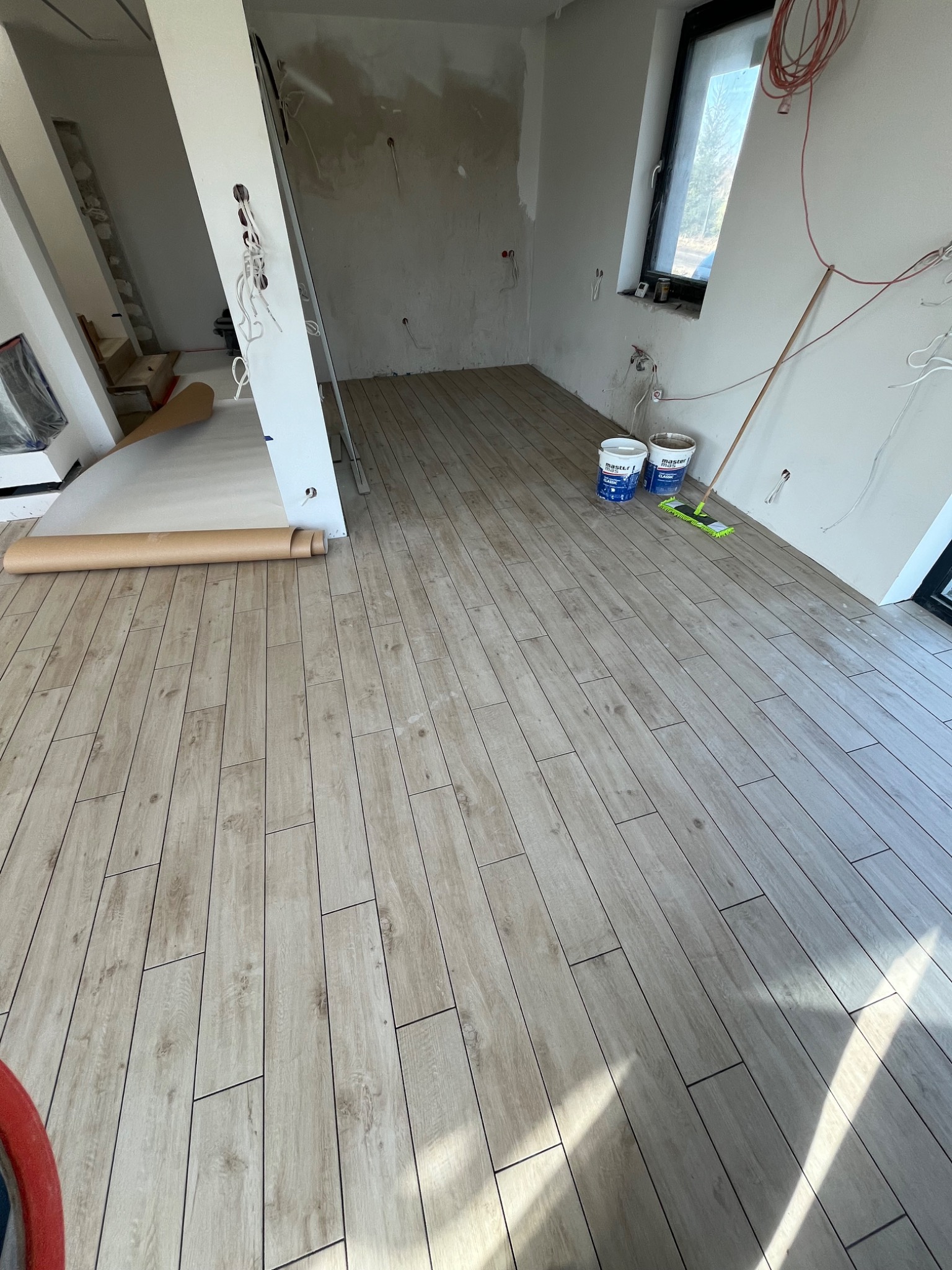 Wnętrze w trakcie remontu z ułożoną podłogą imitującą drewno, widoczne dwa wiadra z farbą, mop, drabina, przygotowana ściana do malowania oraz zwinięta folia malarska.