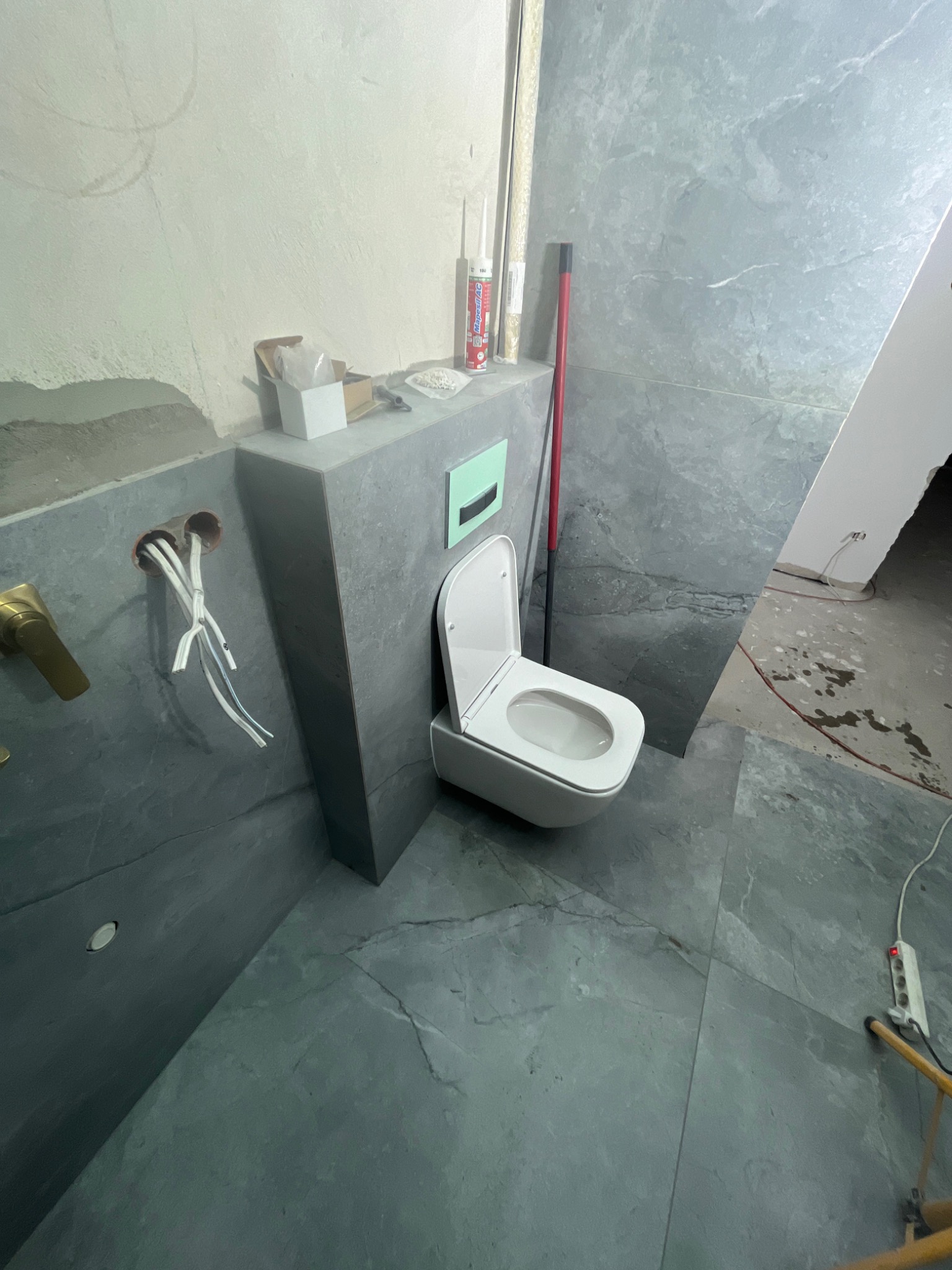 Montaż wiszącej toalety w łazience wykończonej szarymi płytkami imitującymi kamień, widoczne wystające przewody elektryczne i akcesoria montażowe.