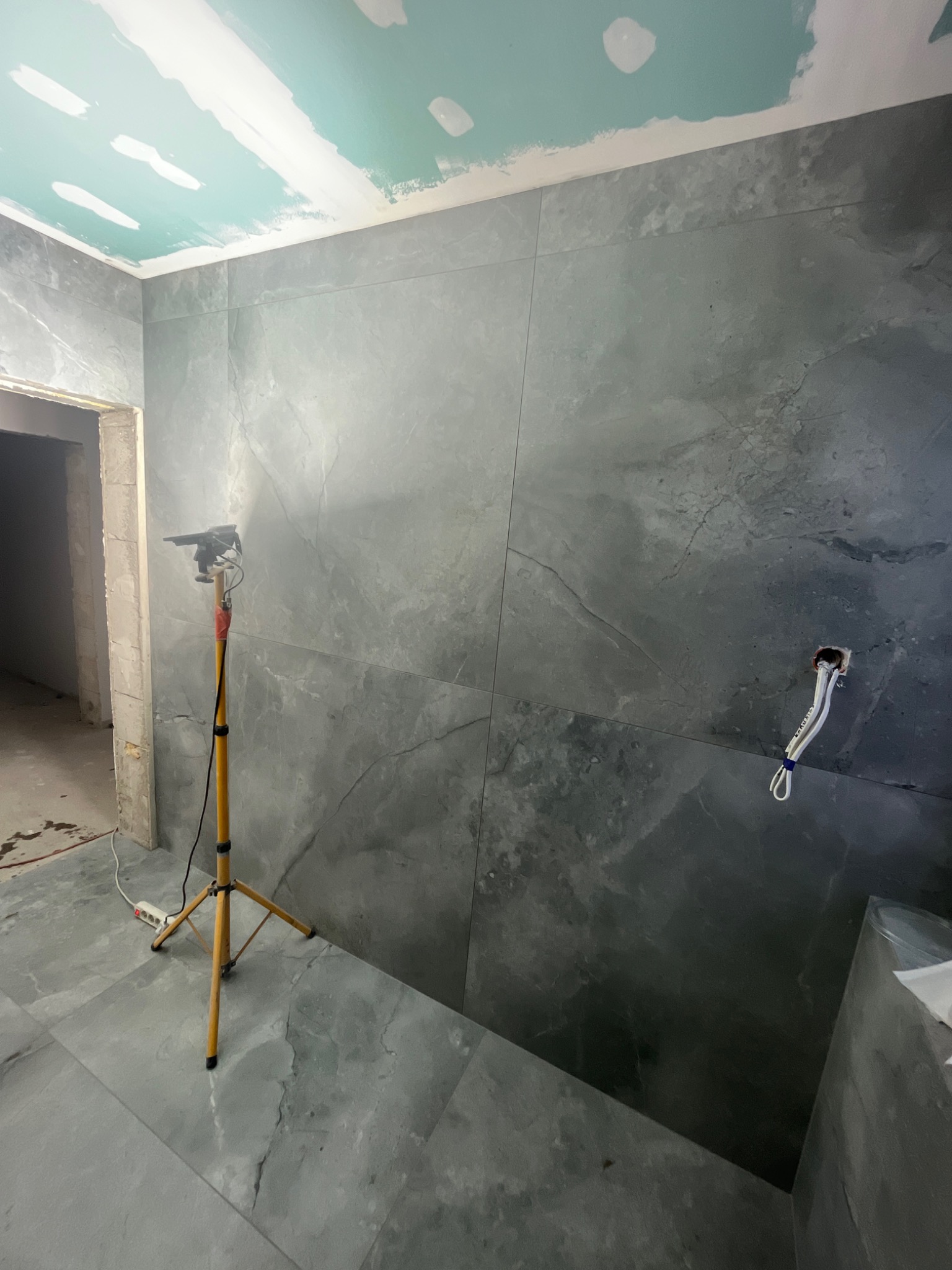 Ściana i podłoga wyłożone szarymi płytkami imitującymi marmur, sufit w trakcie wykańczania płytami gipsowo-kartonowymi, widoczna lampa budowlana na trójnogu i wystające kable elektryczne.