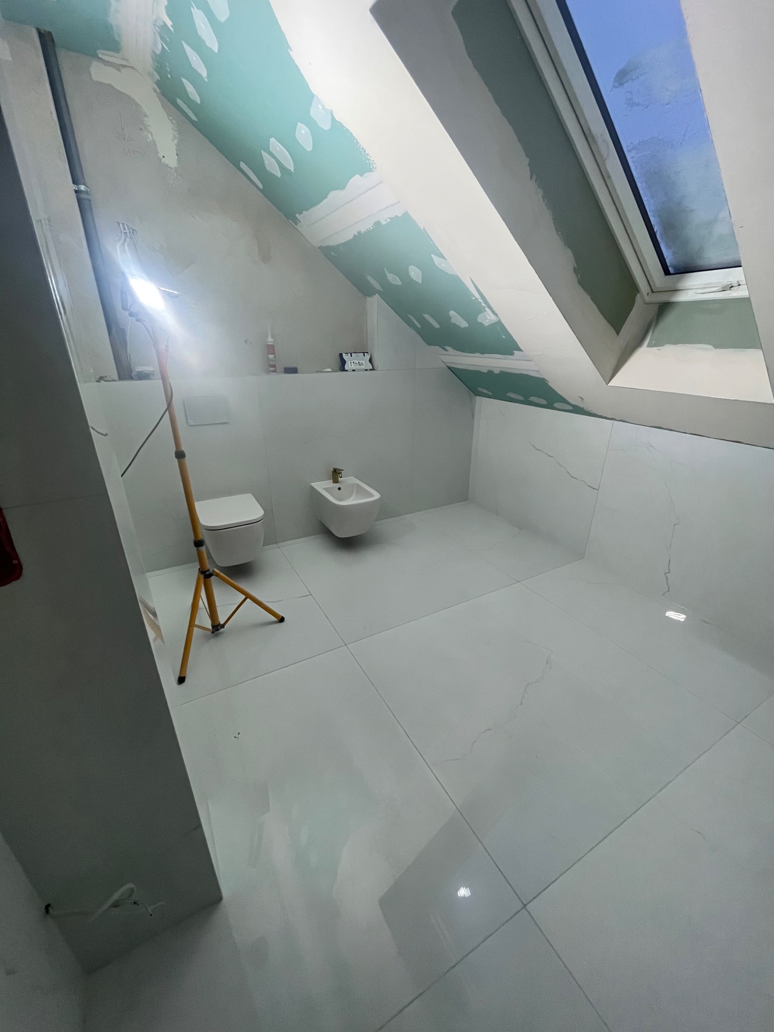 Łazienka na poddaszu w trakcie wykańczania, widoczne białe płytki imitujące marmur, zamontowana toaleta i bidet, sufit skośny z oknem dachowym, częściowo pokryty płytami kartonowo-gipsowymi...
