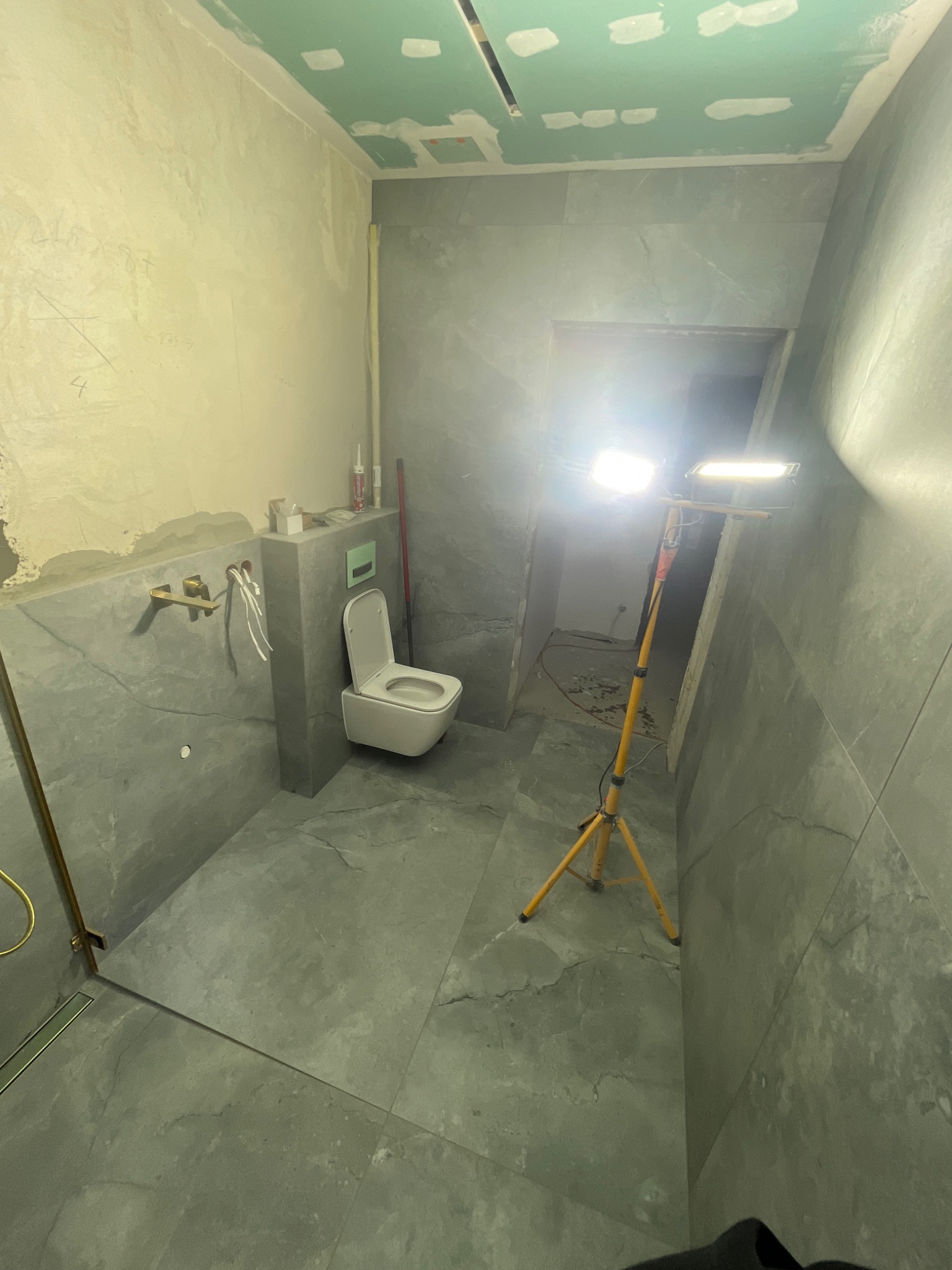 Wnętrze łazienki w trakcie remontu, z szarymi płytkami imitującymi kamień, podwieszaną toaletą, widocznymi instalacjami hydraulicznymi, niedokończoną ścianą z widocznymi śladami po szpachlowaniu...