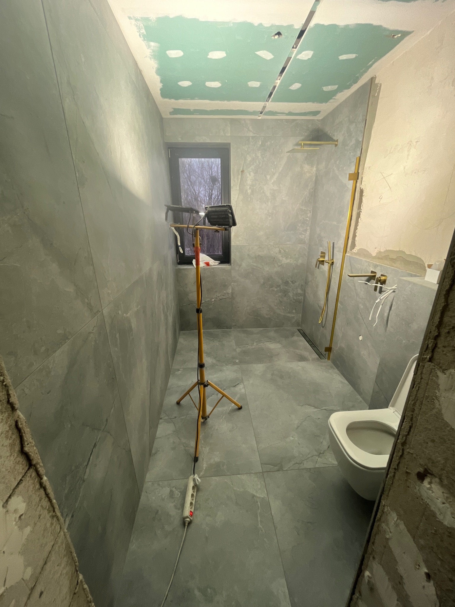 Łazienka w trakcie remontu: szare płytki na ścianach i podłodze, częściowo wykończony sufit z płyt karton gips, złota armatura, oświetlenie robocze na statywie, widoczny fragment toalety...