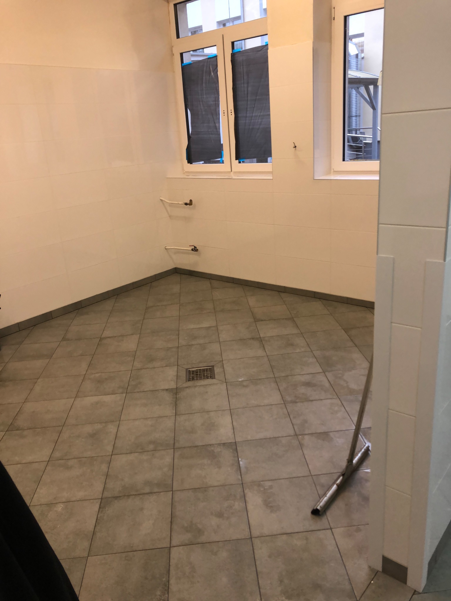 Wykończona łazienka z jasnymi płytkami na ścianach i podłodze. Widoczne okna z zasłoniętymi szybami i metalowy mop przy ścianie. Minimalistyczny styl.