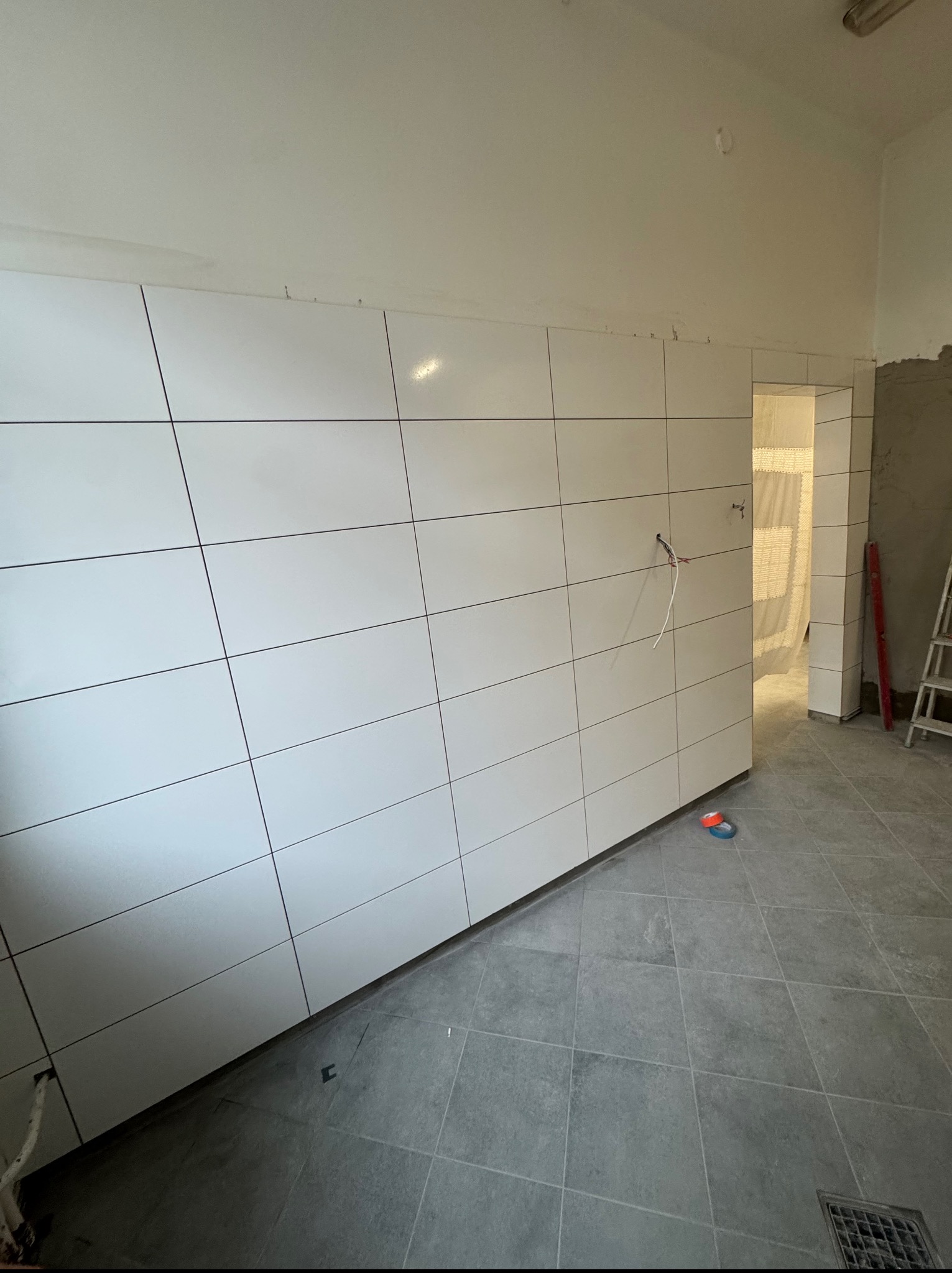 Białe, prostokątne płytki ścienne z ciemnymi fugami w łazience, szare płytki podłogowe. Widoczne przewody elektryczne i poziomica. Częściowo wykończone wnętrze.