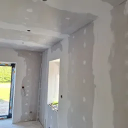 Wnętrze w trakcie prac wykończeniowych: ściany i sufit pokryte gładzią szpachlową, widoczne otwory na oświetlenie, okno i drzwi balkonowe. Na podłodze resztki materiałów.