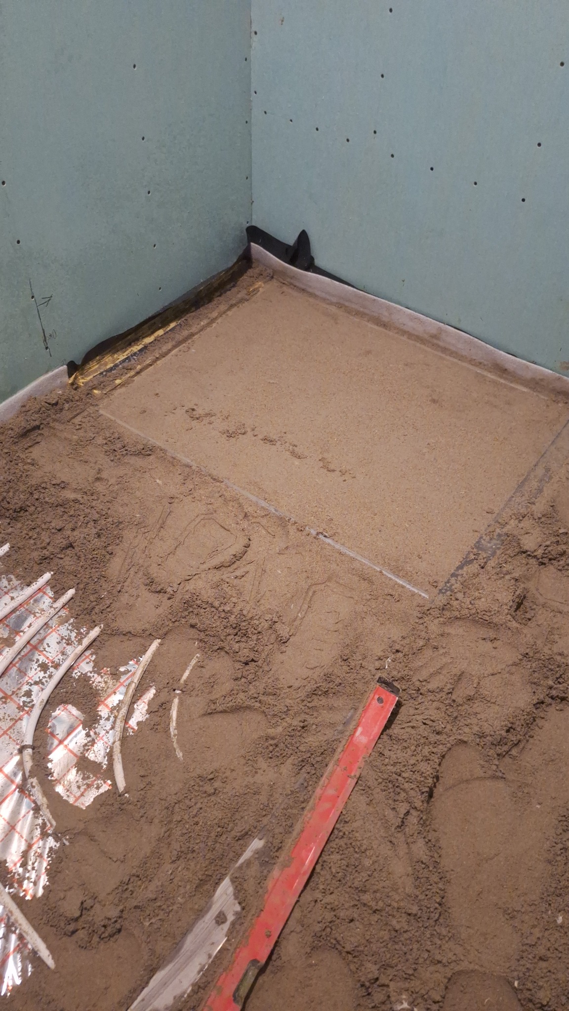 Przygotowanie podłoża pod posadzkę: rury ogrzewania podłogowego częściowo zasypane piaskiem, widoczna poziomica i ściany z płyty karton-gips.