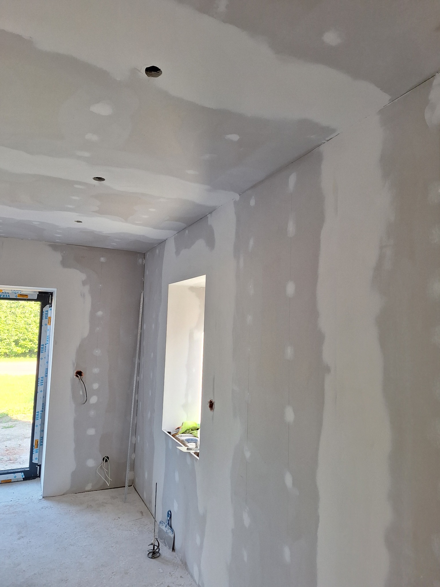 Wnętrze w trakcie prac wykończeniowych: ściany i sufit pokryte gładzią szpachlową, widoczne otwory na oświetlenie, okno i drzwi balkonowe. Na podłodze resztki materiałów.