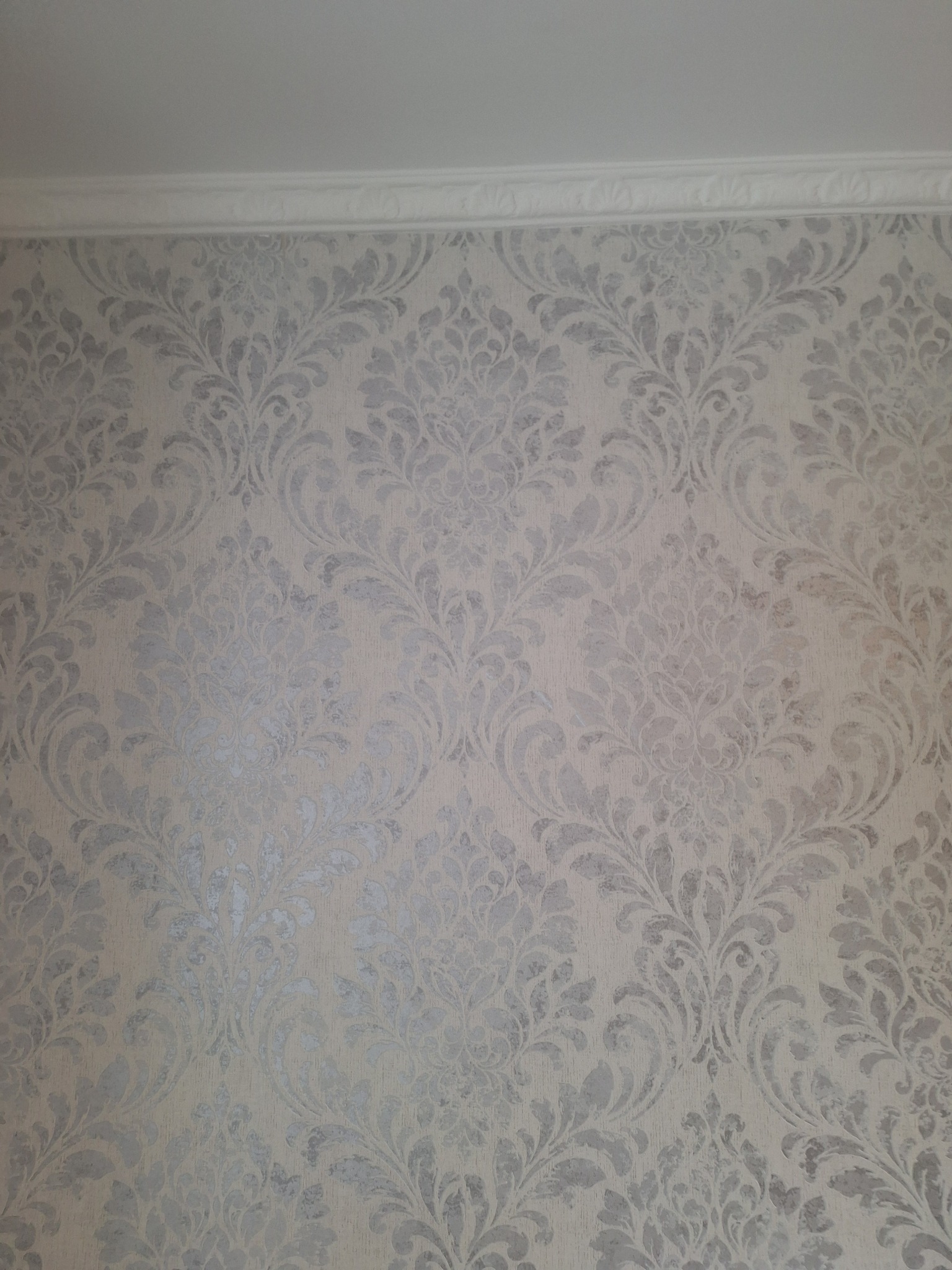 Ściana z beżową tapetą w srebrne, roślinne ornamenty, wykończona białym, ozdobnym gzymsem sufitowym. Delikatne, stonowane barwy i klasyczny wzór.