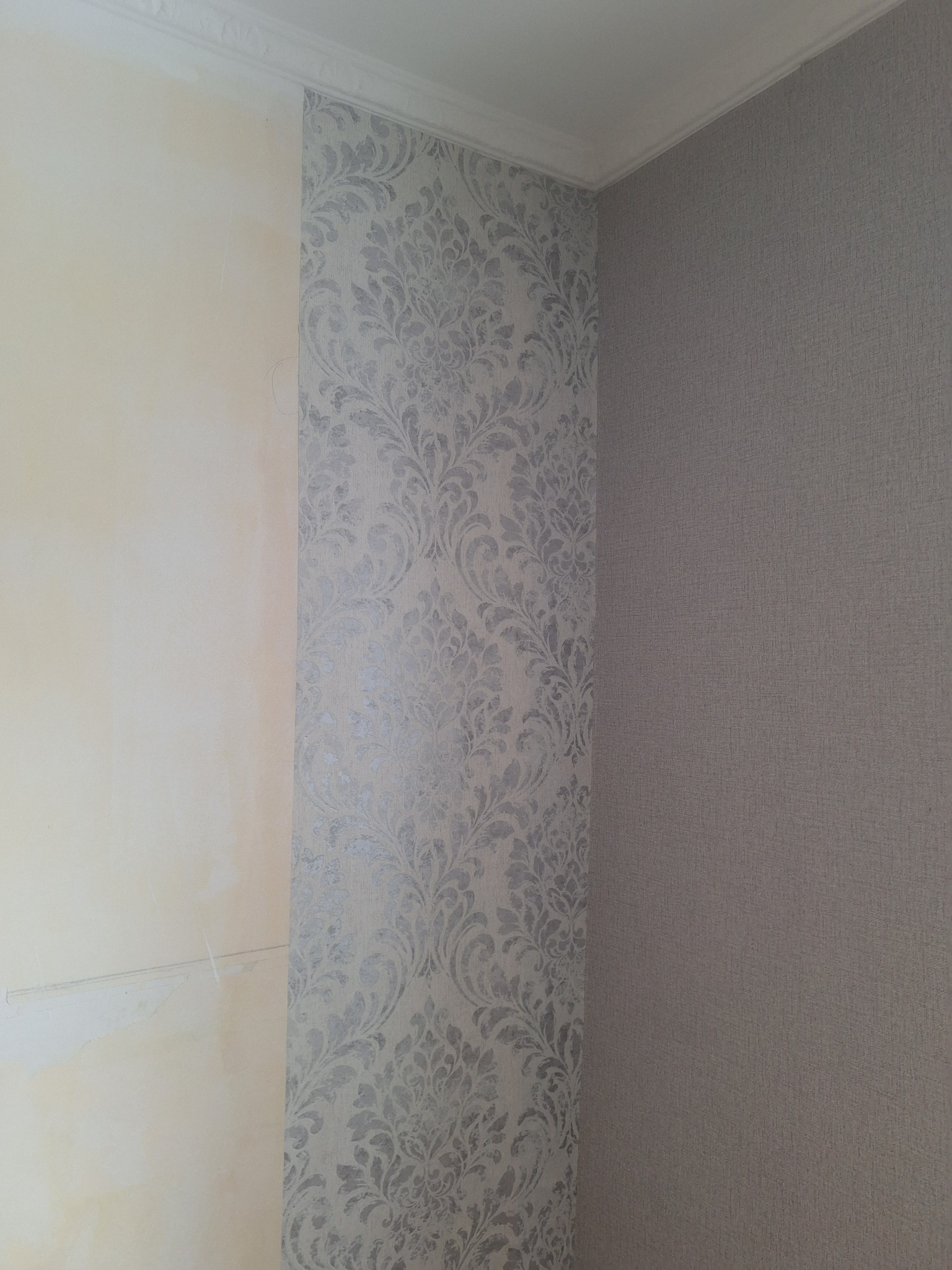 Fragment pokoju: ściana z tapetą w srebrne ornamenty, gładka szara ściana i fragment ściany przygotowanej do tapetowania. Widoczny narożnik i listwa przysufitowa.