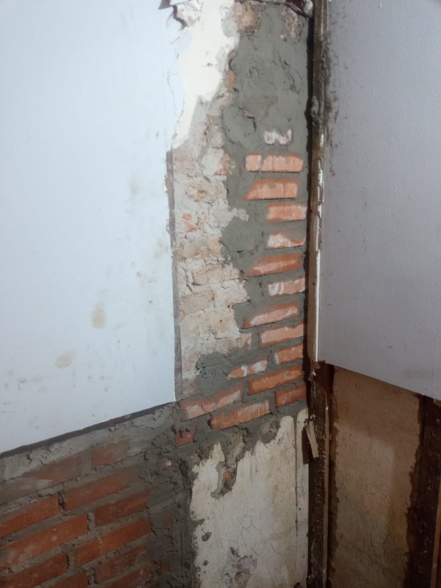 Fragment ściany z odsłoniętą cegłą i świeżym cementem, widoczne ślady po usuniętych płytkach, przygotowanie do dalszych prac wykończeniowych w pomieszczeniu.