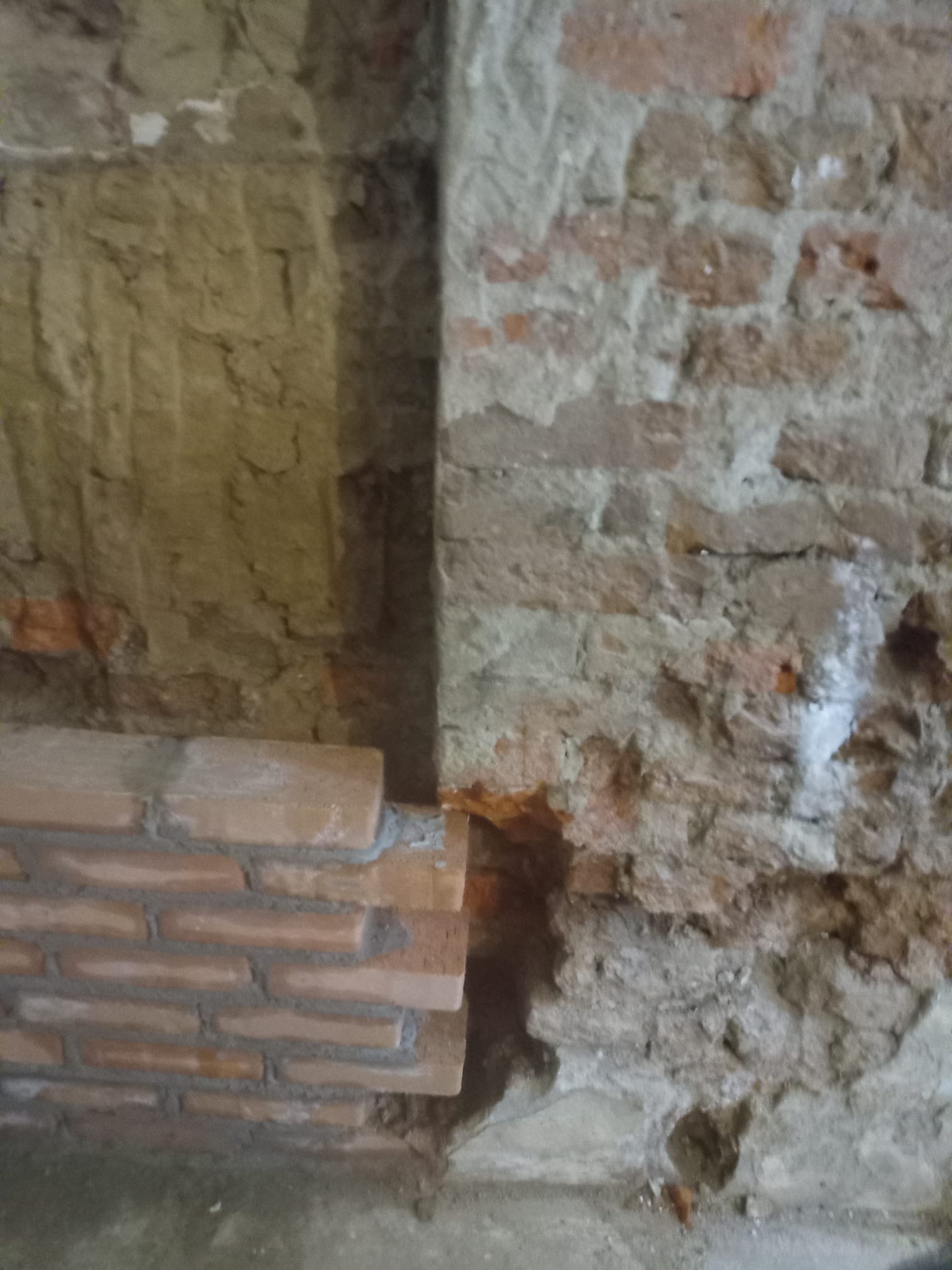 Fragment muru z cegły, częściowo otynkowany, z widocznym nowym fragmentem ceglanej ściany w trakcie budowy. Surowe wykończenie, detale spoin i faktury materiałów.