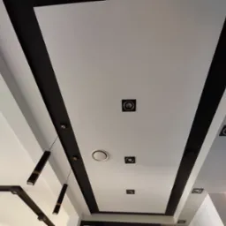 Nowoczesny sufit z białymi panelami, czarnymi pasami dekoracyjnymi, punktowymi lampami i wiszącym oświetleniem.