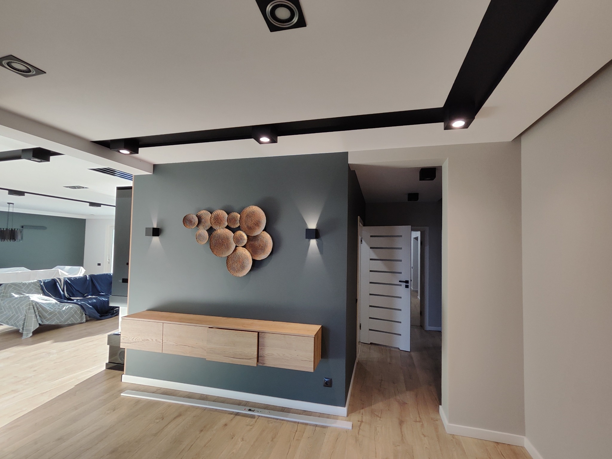 Nowoczesne wnętrze po remoncie: szara ściana z dekoracją, drewniana szafka RTV, podłoga panelowa, białe drzwi z poziomymi paskami i oświetlenie punktowe.