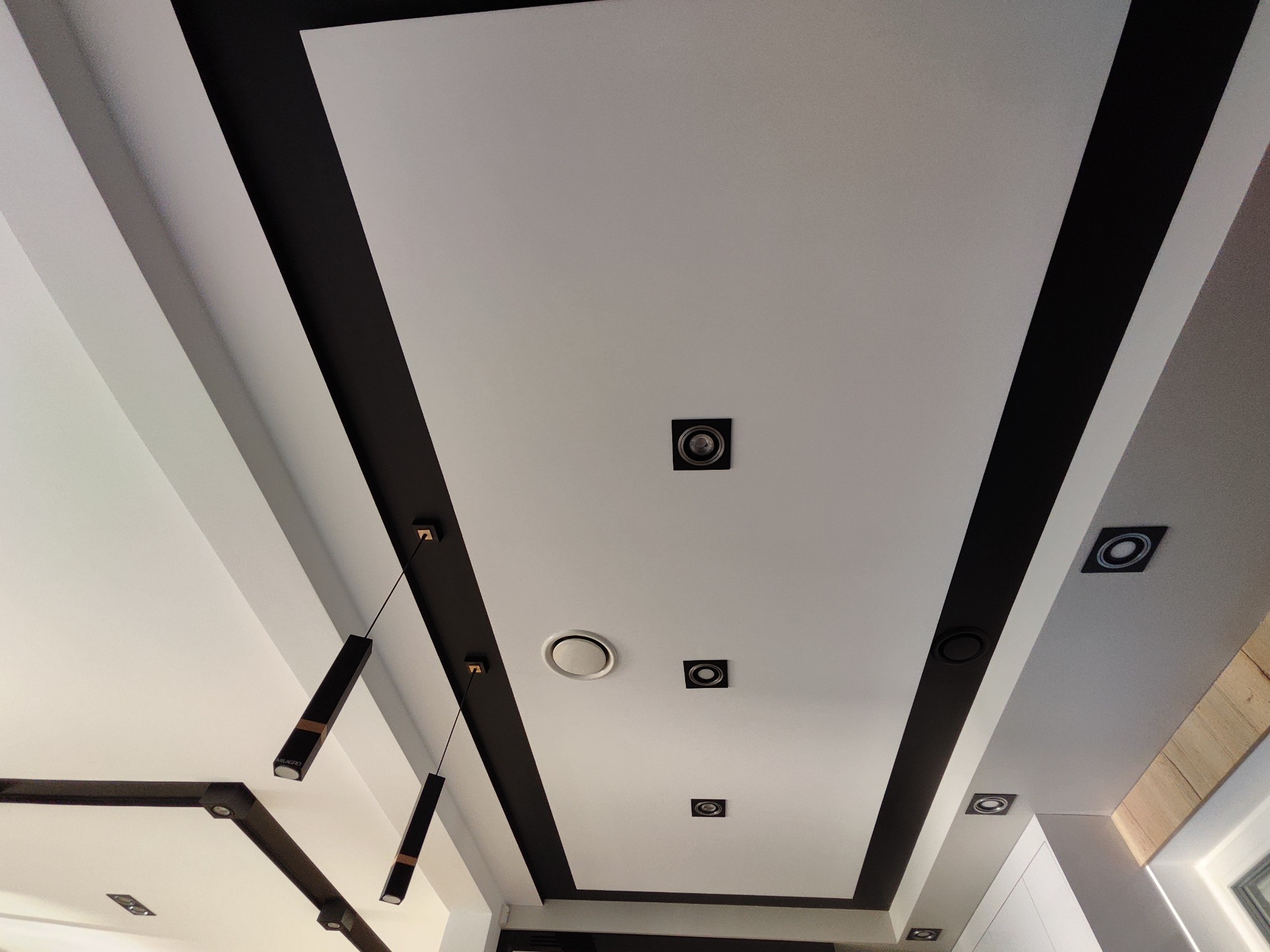 Nowoczesny sufit z białymi panelami, czarnymi pasami dekoracyjnymi, punktowymi lampami i wiszącym oświetleniem.