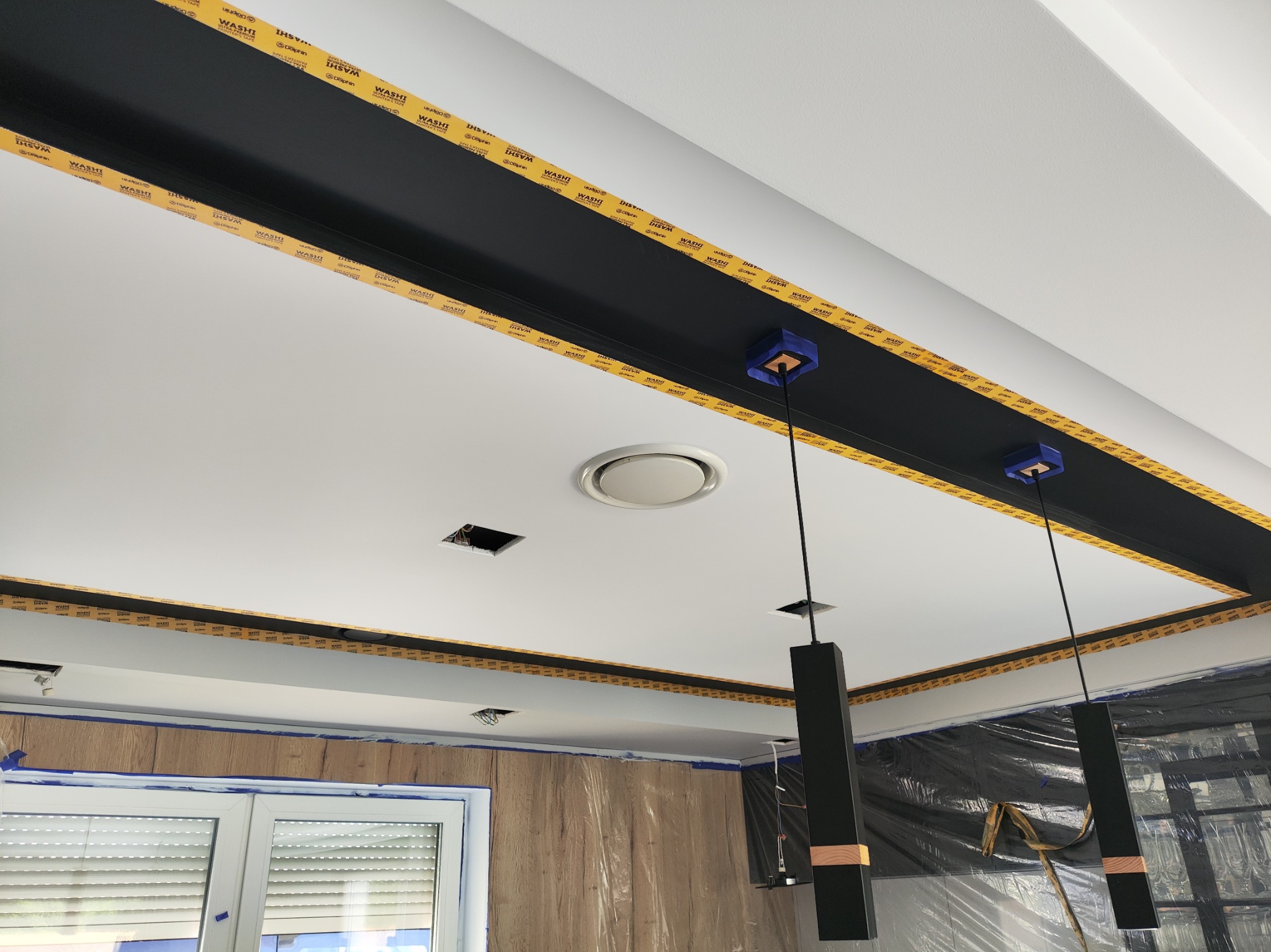 Przygotowanie sufitu do malowania: widoczna czarna wnęka oddzielona taśmą malarską, zabezpieczone ściany folią, wiszące lampy i otwory na oświetlenie.