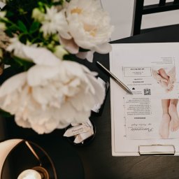 Pracownia Zdrowia i Urody - Eleganckie biurko z dokumentami dotyczącymi podologii, ozdobione białymi piwoniami w wazonie i designerską lampą.