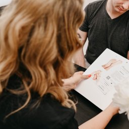 Pracownia Zdrowia i Urody - Klient w salonie podologicznym przegląda ofertę zabiegów na stopy, prezentowaną na karcie informacyjnej przez specjalistkę z blond włosami, w tle bukiet białych kwiatów.