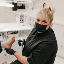 Pracownia Zdrowia i Urody - Specjalistka w czarnym uniformie i masce ochronnej, w trakcie zabiegu pedicure frezarką, skupiona na stopie klienta, w tle białe meble i lampa kosmetyczna.