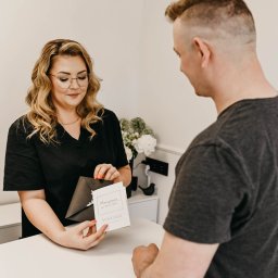 Pracownia Zdrowia i Urody - Recepcjonistka w czarnym uniformie wręcza klientowi voucher prezentowy w eleganckiej kopercie w salonie kosmetycznym z białym biurkiem i kwiatami w tle.