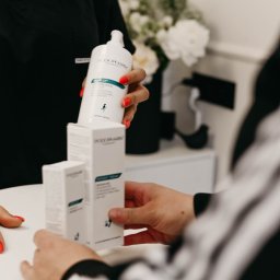 Pracownia Zdrowia i Urody - Prezentacja zestawu kosmetyków Podopharm: butelka Sunflex dermatological body soap oraz pudełka Oxygen Cream, na białym stole w gabinecie kosmetycznym, z widocznymi dłońmi personelu i klienta.