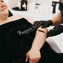 Pracownia Zdrowia i Urody - Kobieta poddaje się zabiegowi pijawkami na ramieniu w gabinecie, terapeutka w czarnych rękawiczkach przykłada pijawkę, pacjentka leży pod czarnym welurowym okryciem z logo salonu, w tle świece...