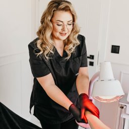 Pracownia Zdrowia i Urody - Kosmetolog w czarnym uniformie i rękawiczkach wykonuje zabieg na dłoni klienta pod lampą emitującą czerwone światło.