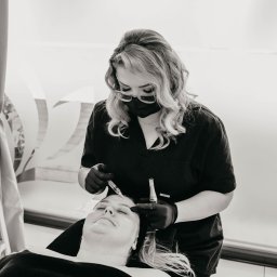 Pracownia Zdrowia i Urody - Czarno-białe zdjęcie z bliska: kosmetolog w czarnej masce i rękawiczkach wykonuje zabieg na twarzy klientki leżącej na fotelu, używając strzykawki z igłą; widoczny napis 'Pracownia Zdrowia i Urody'...