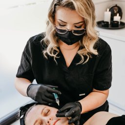 Pracownia Zdrowia i Urody - Kobieta w czarnym uniformie i rękawiczkach wykonuje iniekcję w policzek pacjentki leżącej na leżance, w tle widoczne świece i dekoracje.