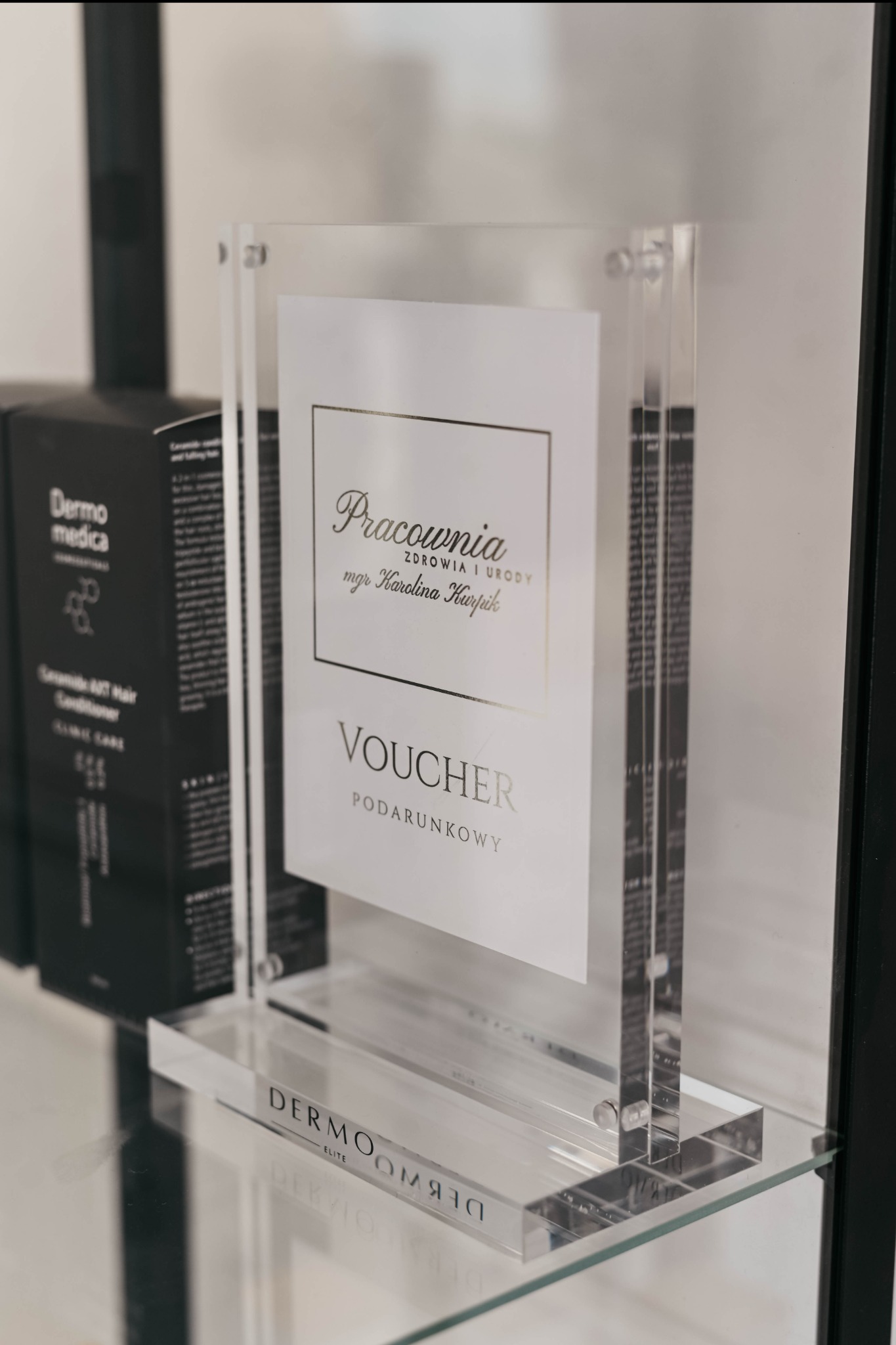 Przezroczysty stojak reklamowy z voucherem prezentowym 'Pracownia Zdrowia i Urody mgr Karolina Hupik' na szklanej półce, obok kosmetyki Dermo medica.