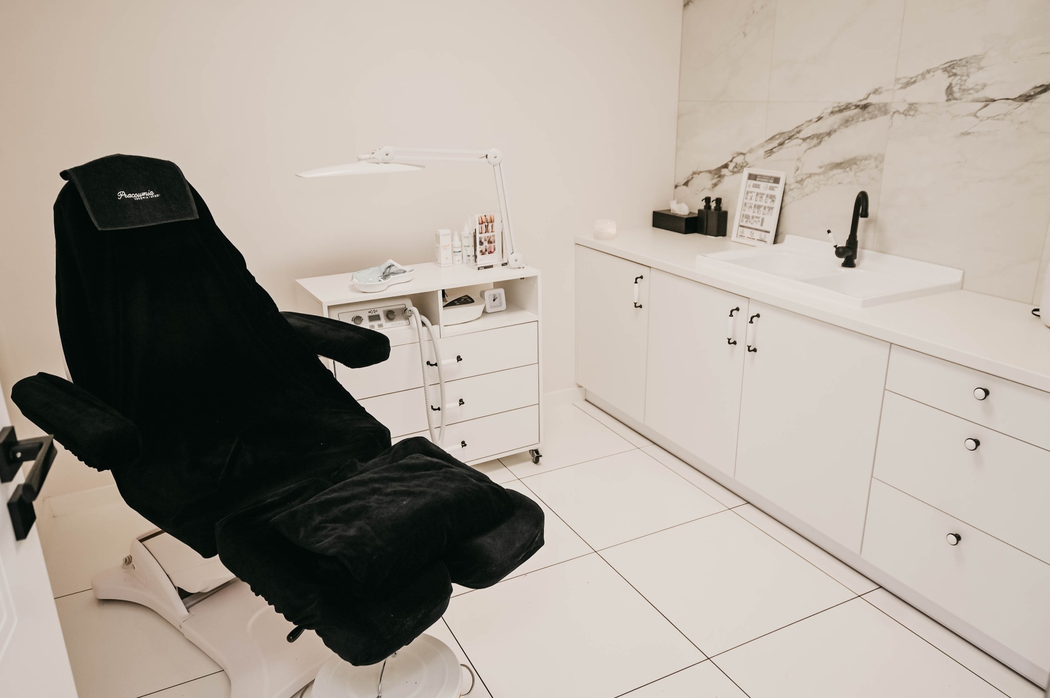 Nowoczesny gabinet kosmetyczny z czarnym, welurowym fotelem zabiegowym, białymi meblami i marmurową ścianą w tle.