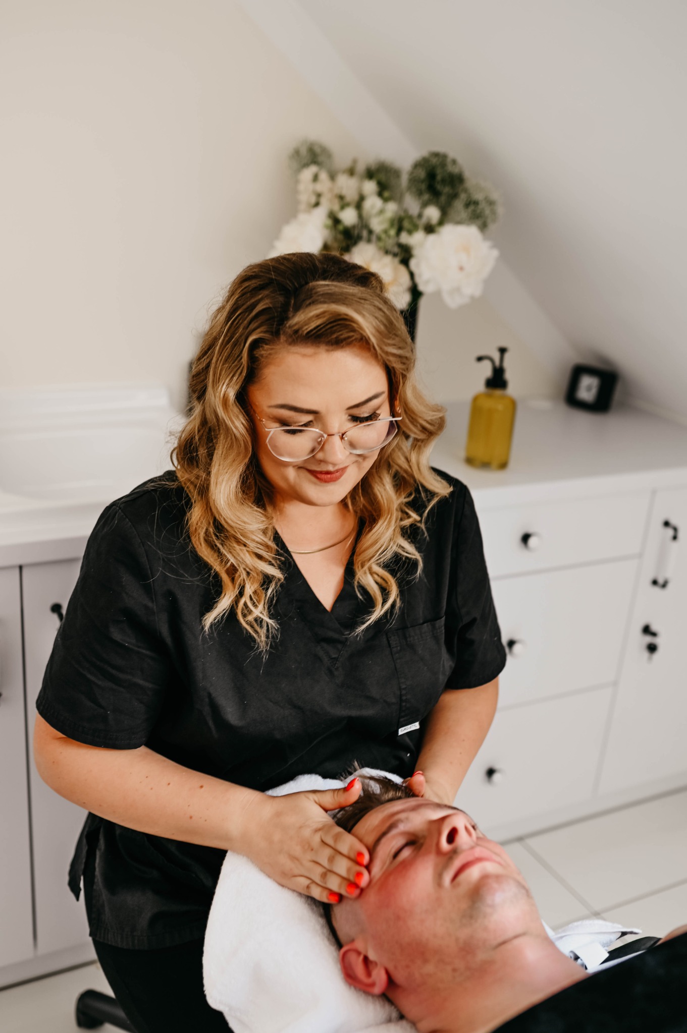 Kobieta w czarnym uniformie i okularach wykonuje masaż twarzy mężczyźnie leżącemu na białym ręczniku; widoczne białe kwiaty w wazonie i biała szafka w tle.