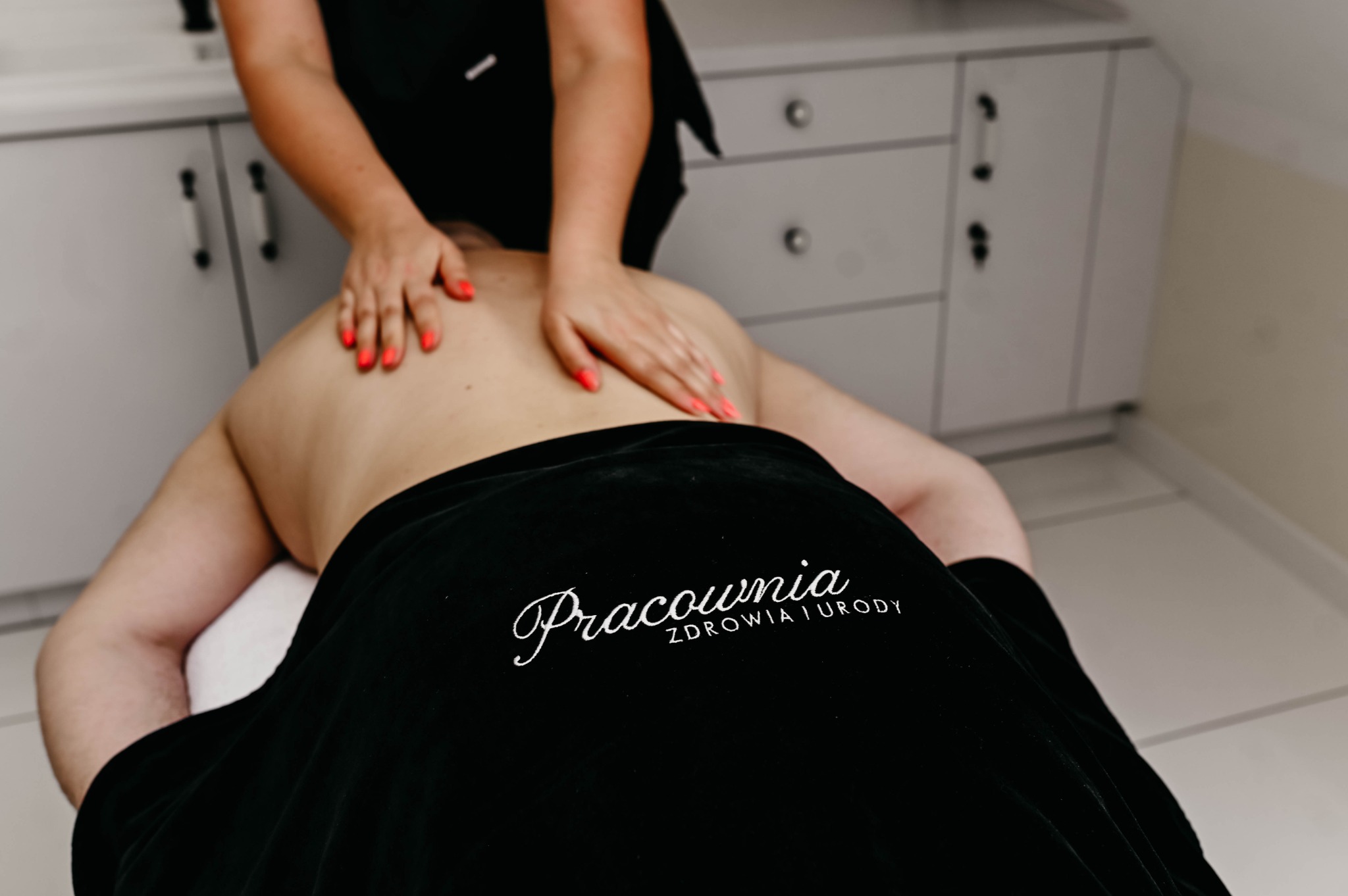 Masaż pleców wykonywany w salonie w Strzałkowie, klientka przykryta czarnym ręcznikiem z logo 'Pracownia Zdrowia i Urody', widoczne dłonie masażystki z czerwonym manicure.