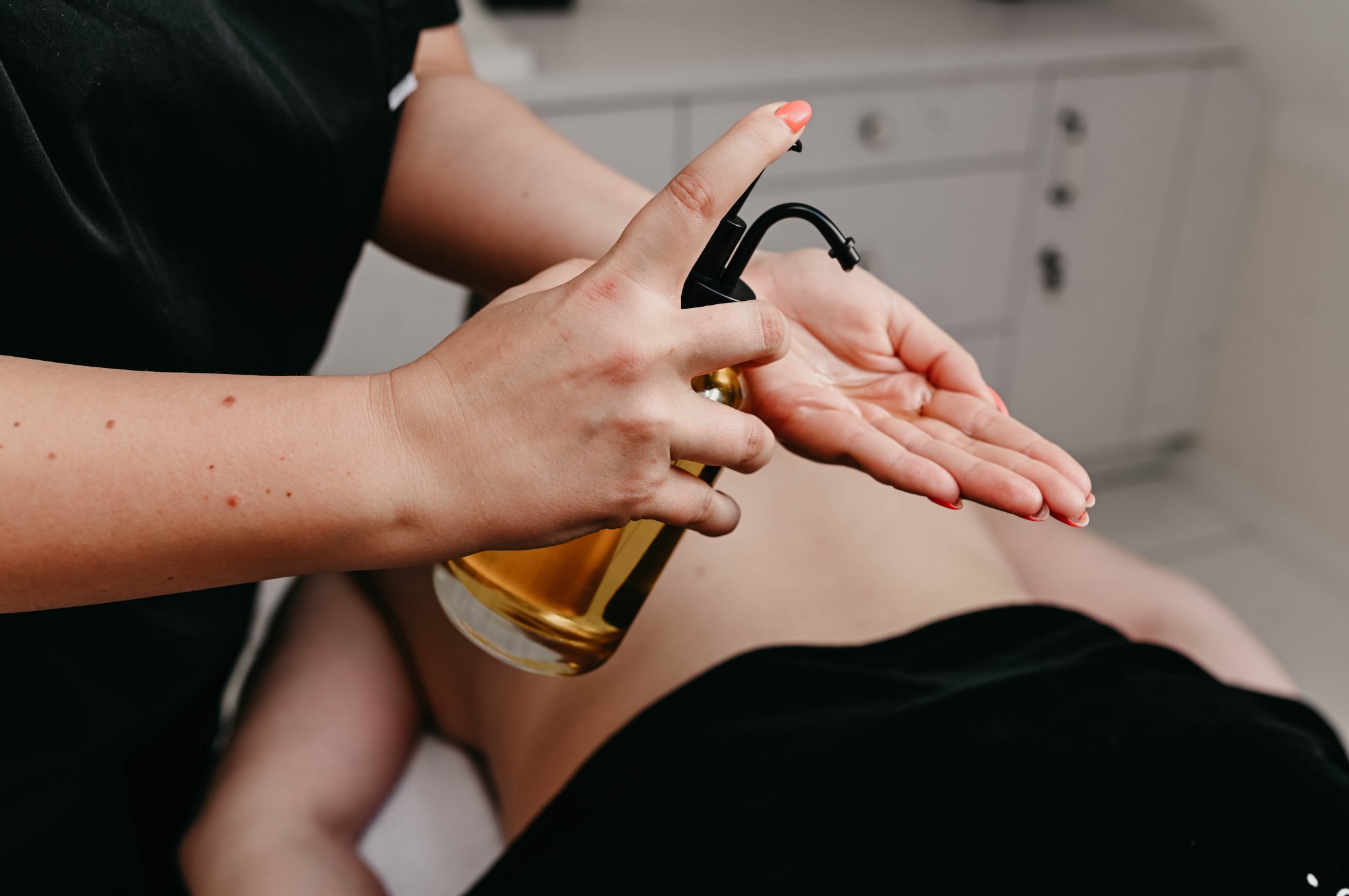 Olej do masażu aplikowany na dłoń przed zabiegiem na ciało; widoczny fragment ciała klienta pod czarnym okryciem, biała szafka w tle.