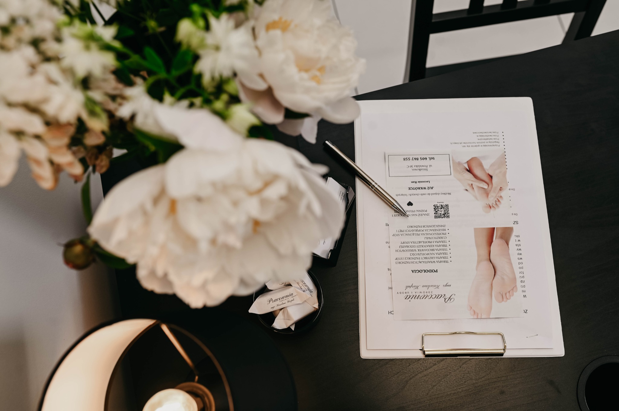 Eleganckie biurko z dokumentami dotyczącymi podologii, ozdobione białymi piwoniami w wazonie i designerską lampą.