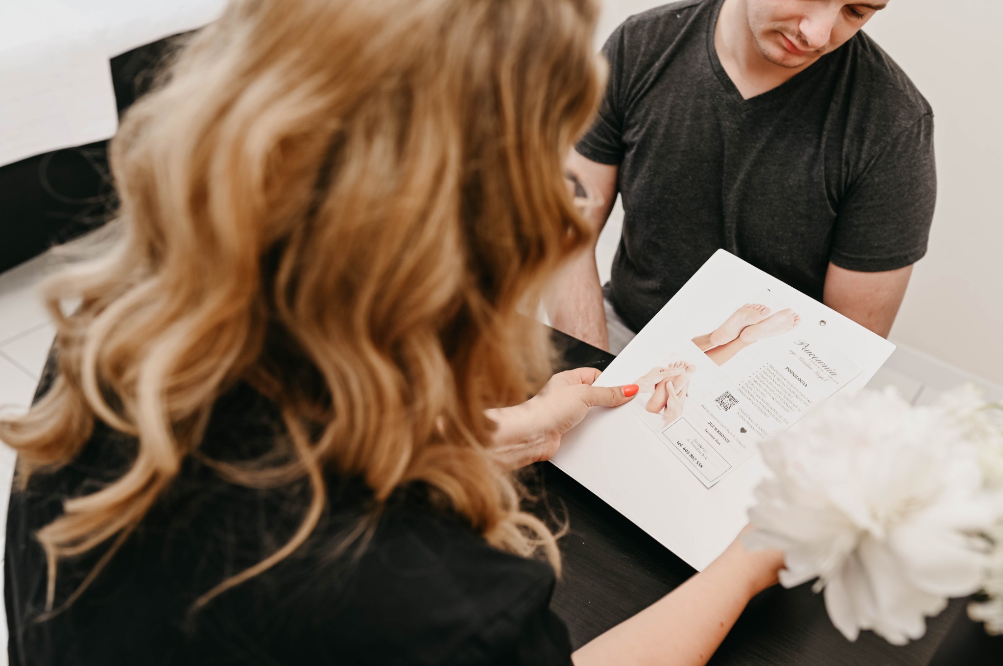 Klient w salonie podologicznym przegląda ofertę zabiegów na stopy, prezentowaną na karcie informacyjnej przez specjalistkę z blond włosami, w tle bukiet białych kwiatów.