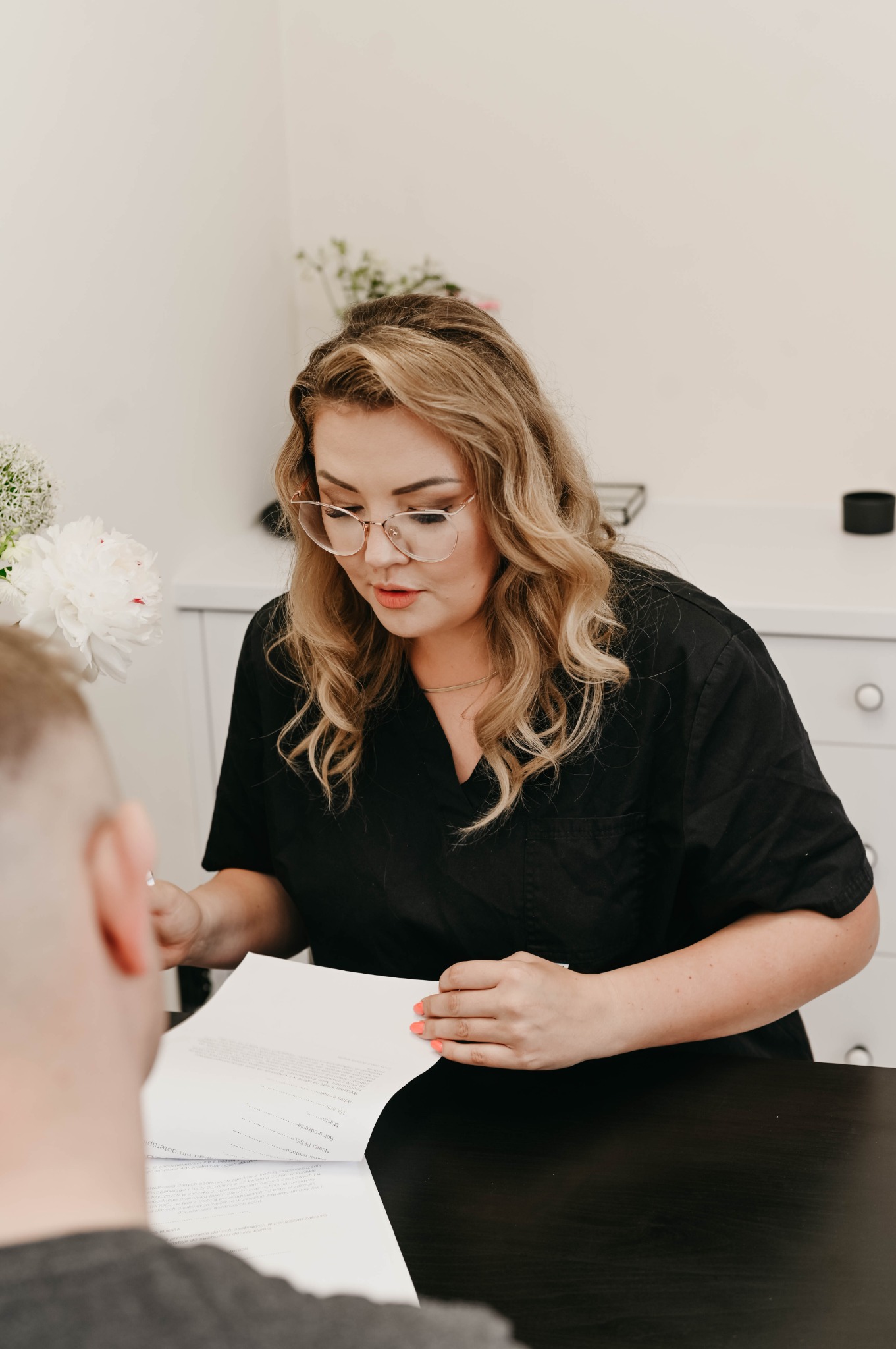 Kobieta w czarnym uniformie i okularach omawia dokumenty z klientem przy czarnym stole, w tle białe kwiaty i szafka.