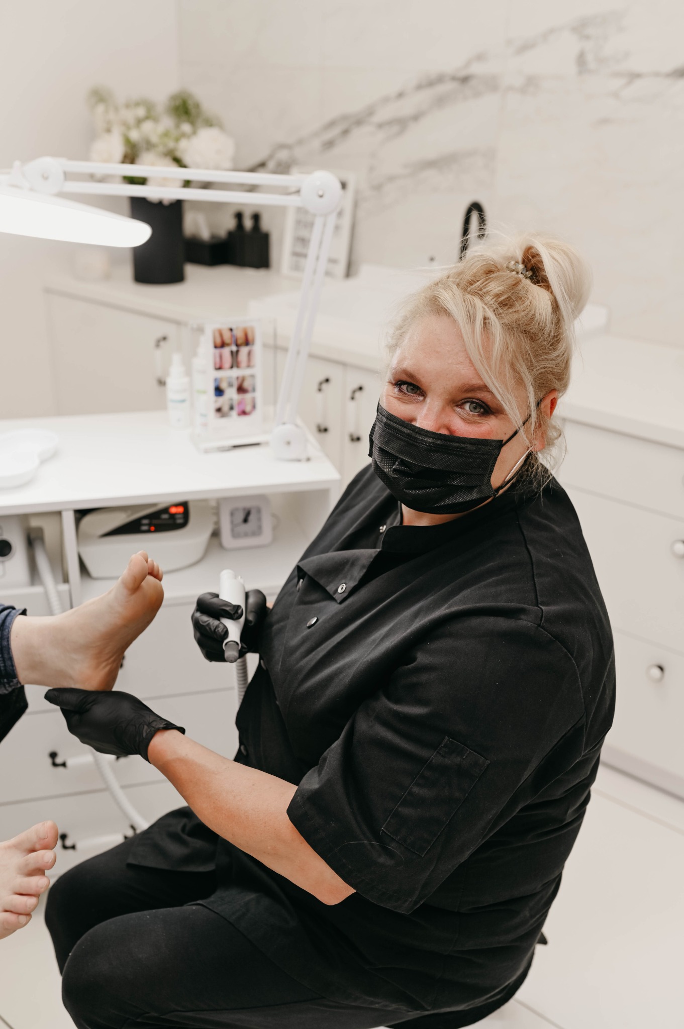 Specjalistka w czarnym uniformie i masce ochronnej, w trakcie zabiegu pedicure frezarką, skupiona na stopie klienta, w tle białe meble i lampa kosmetyczna.