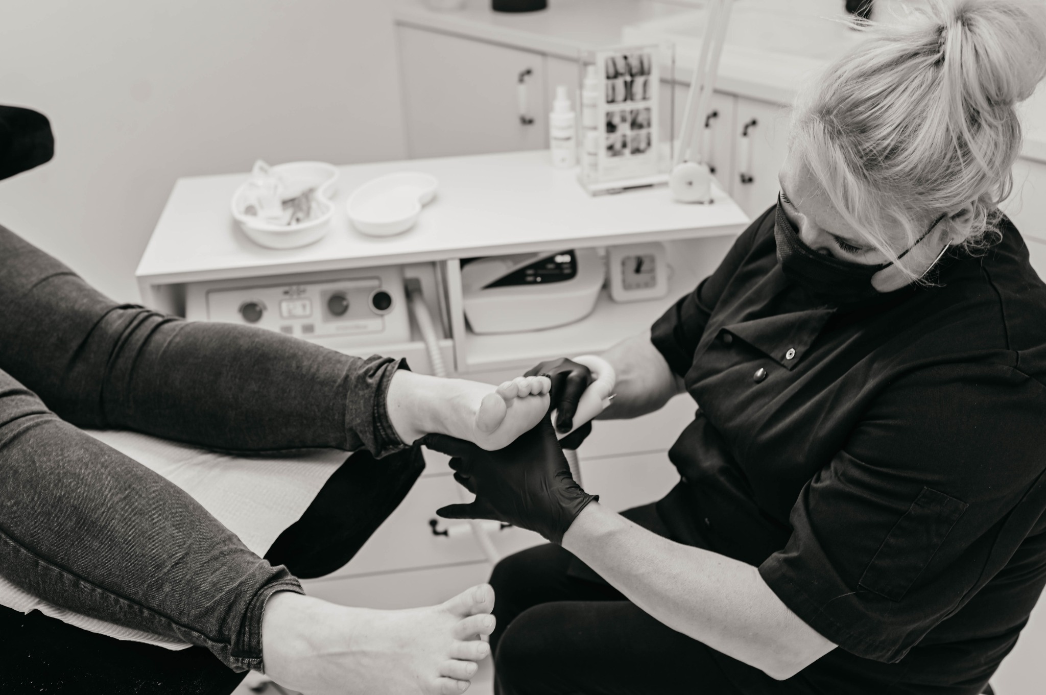 Stylizowana fotografia w odcieniach szarości przedstawia kosmetyczkę w czarnych rękawiczkach, która wykonuje zabieg na stopie klientki leżącej na fotelu; widoczne narzędzia i akcesoria na białym...