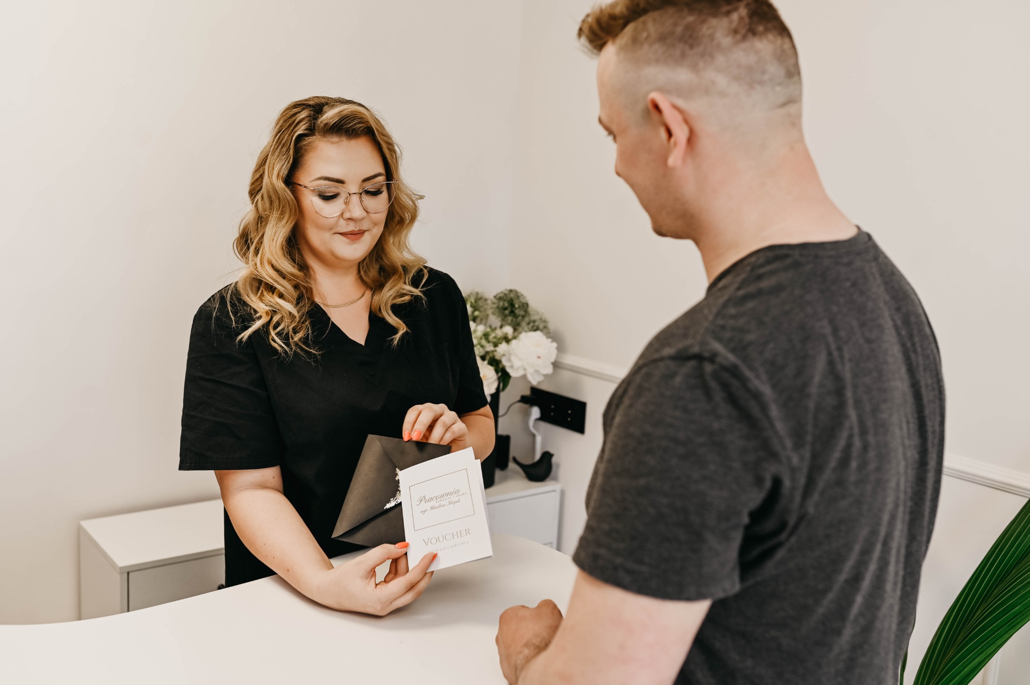 Recepcjonistka w czarnym uniformie wręcza klientowi voucher prezentowy w eleganckiej kopercie w salonie kosmetycznym z białym biurkiem i kwiatami w tle.