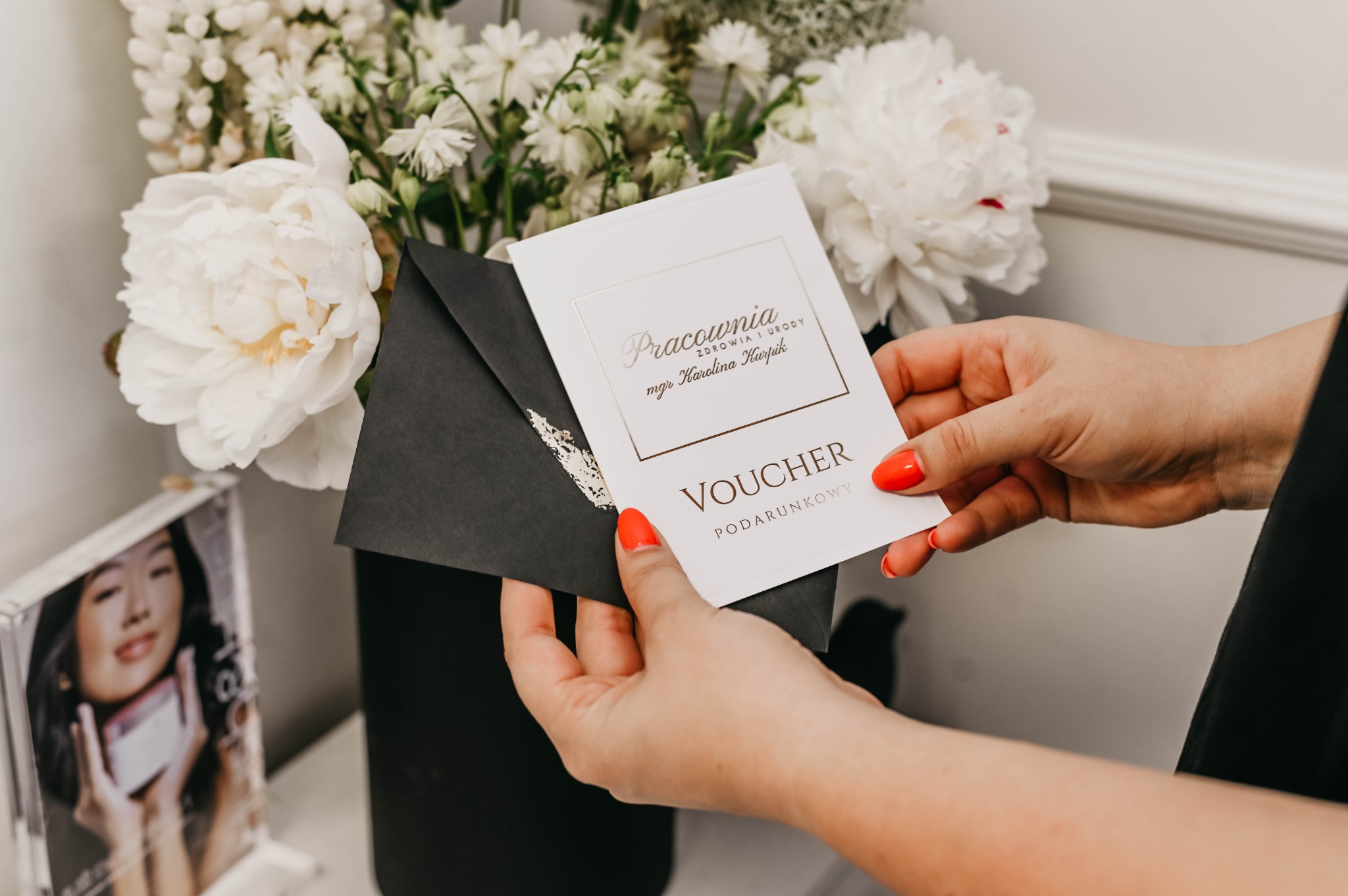 Dłonie z czerwonym manicure trzymają voucher podarunkowy w czarnej kopercie na tle białych kwiatów i zdjęcia kobiety.
