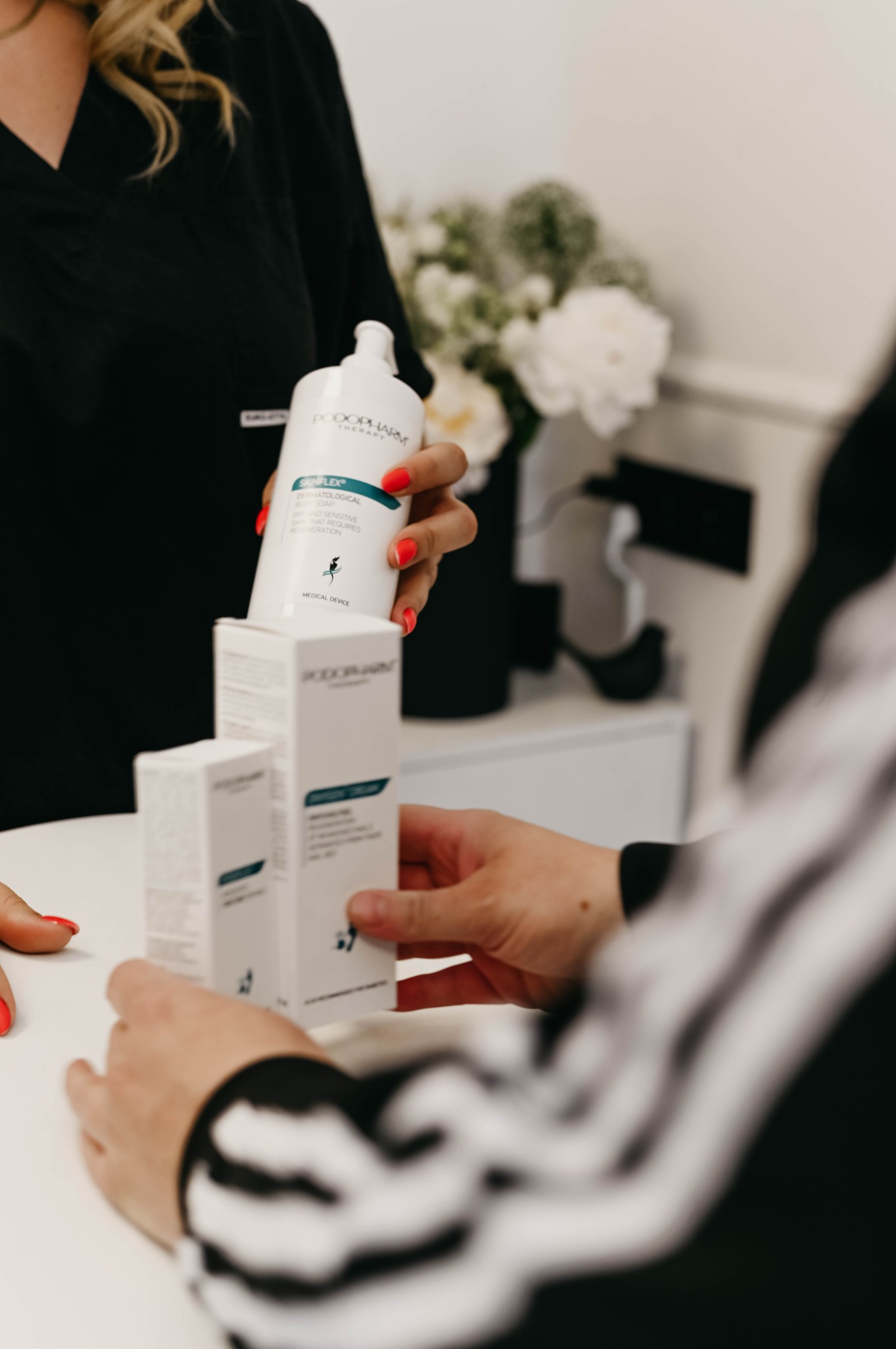 Prezentacja zestawu kosmetyków Podopharm: butelka Sunflex dermatological body soap oraz pudełka Oxygen Cream, na białym stole w gabinecie kosmetycznym, z widocznymi dłońmi personelu i klienta.