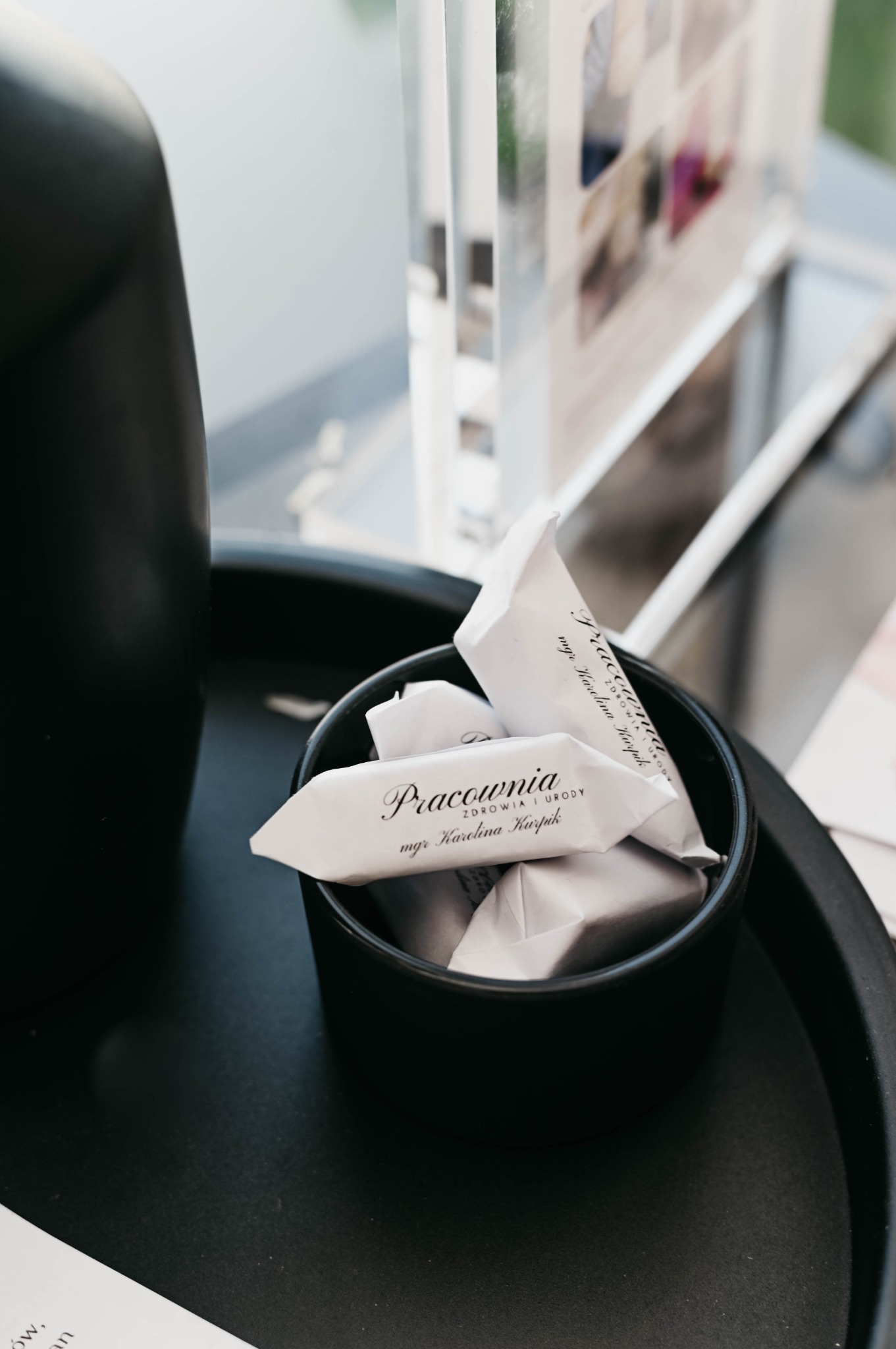 Czarne naczynie wypełnione cukierkami z logo 'Pracownia Zdrowia i Urody mgr Karolina Kurpik', stojące na czarnej tacy obok czarnego wazonu i ulotek reklamowych.