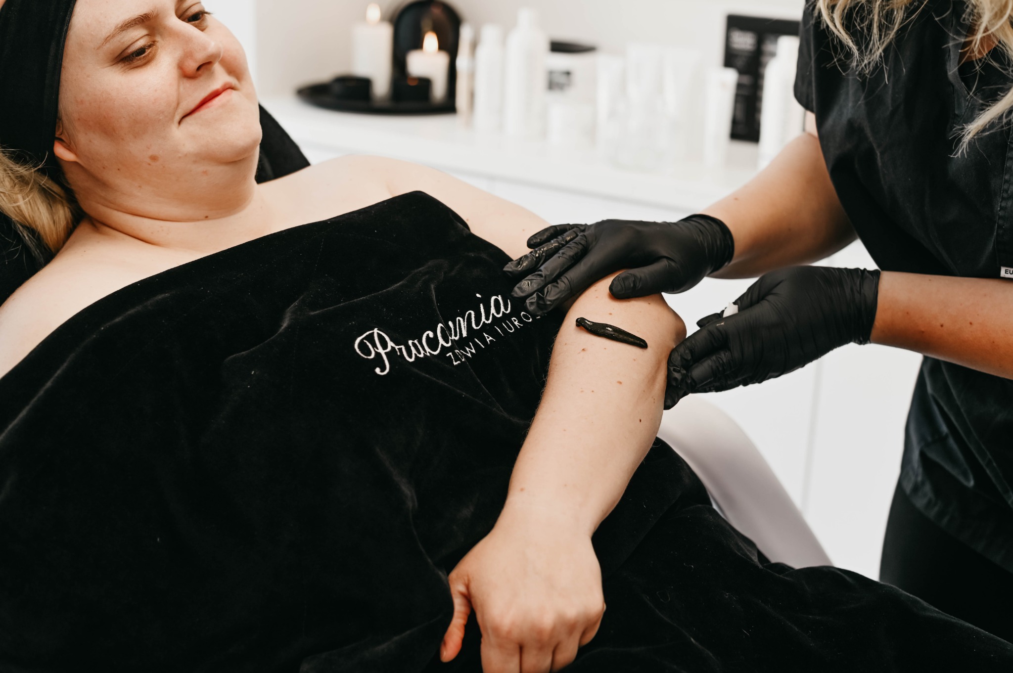 Kobieta poddaje się zabiegowi pijawkami na ramieniu w gabinecie, terapeutka w czarnych rękawiczkach przykłada pijawkę, pacjentka leży pod czarnym welurowym okryciem z logo salonu, w tle świece...