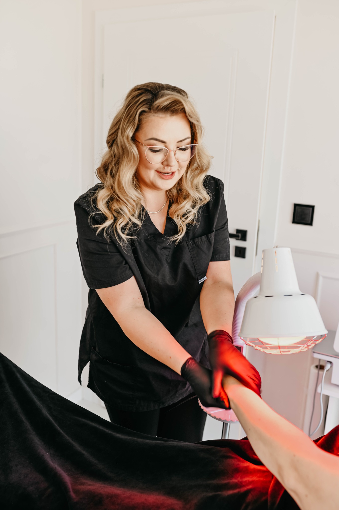 Kosmetolog w czarnym uniformie i rękawiczkach wykonuje zabieg na dłoni klienta pod lampą emitującą czerwone światło.