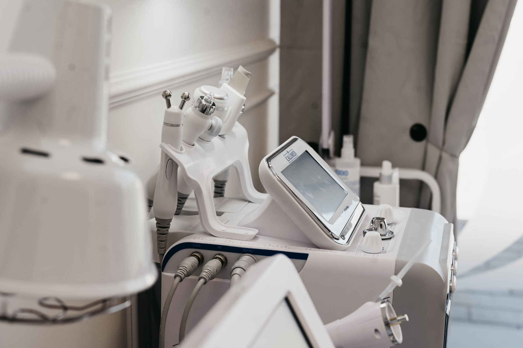 Biały aparat do zabiegów kosmetycznych z różnymi głowicami i ekranem dotykowym, umieszczony w gabinecie z zasłoną w tle.